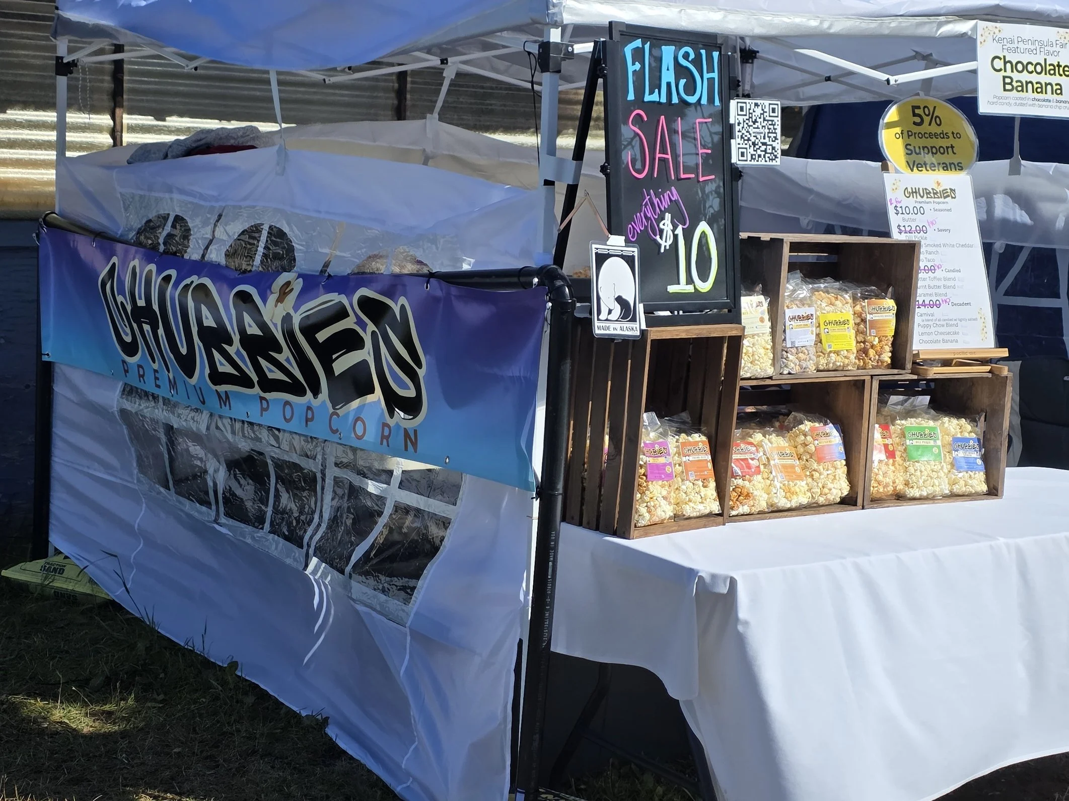 Popcorn stand at an outdoor market. A sign reads 'Flash Sale Everything $10'. Packaged popcorn is displayed in wooden crates. Additional signs advertise product prices and support for veterans.