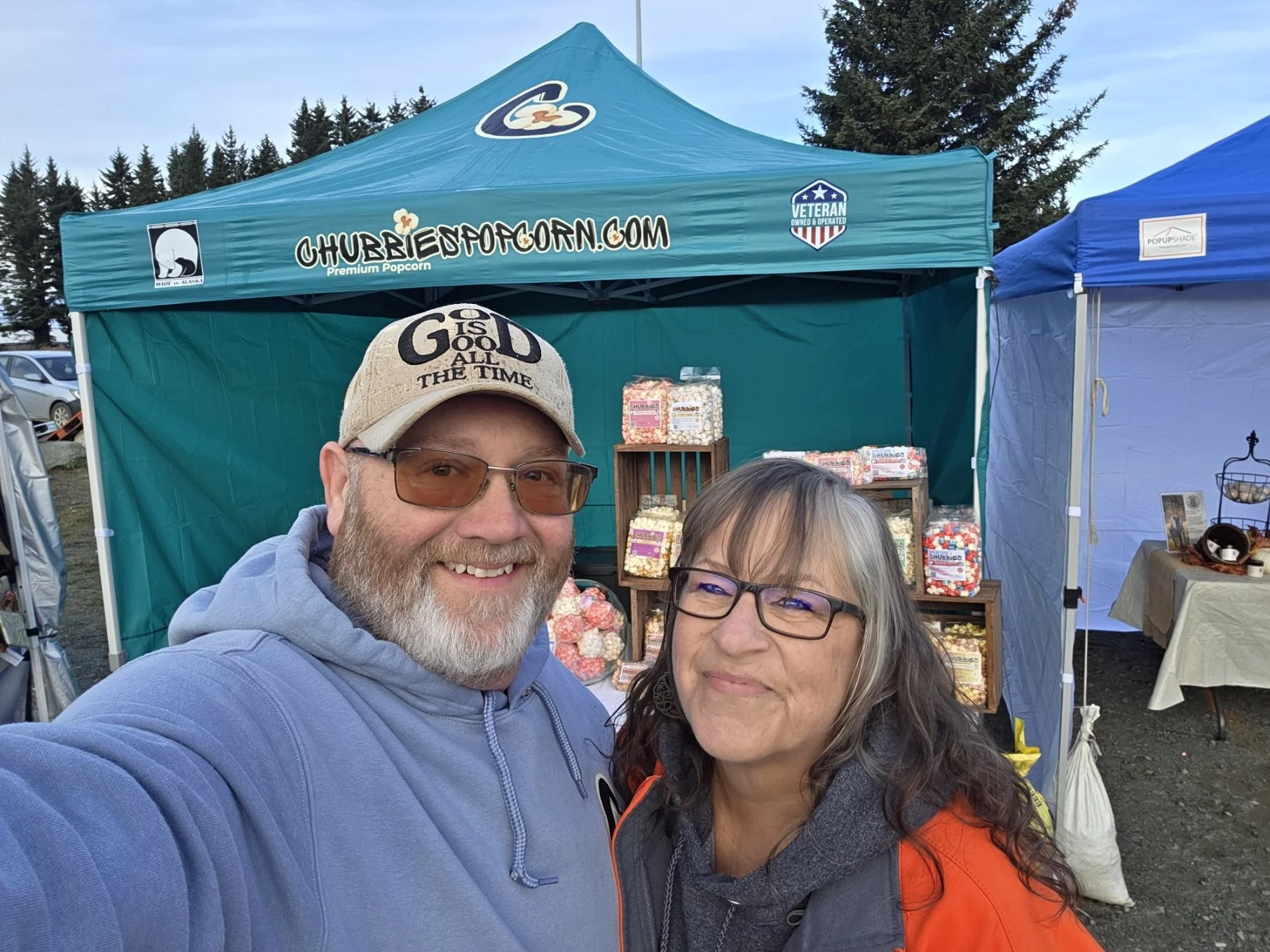 A man and a woman taking a selfie in front of a popcorn stand at an outdoor market or festival with tents and trees in the background.