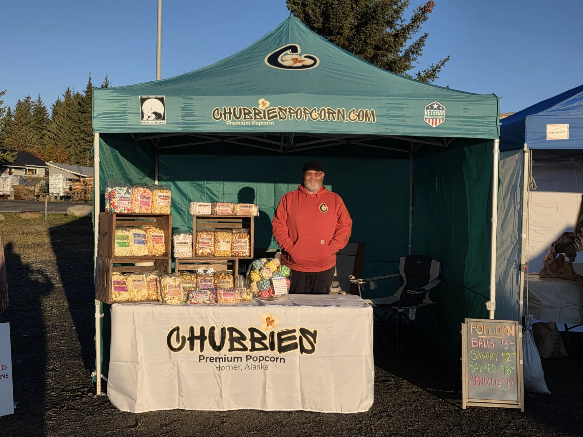 A man wearing a red hoodie standing behind a popcorn stand at an outdoor market. The stand has a green canopy with branding for ChubiesPopcorn.com and a banner with the word 'Chubies' and 'Premium Popcorn' written on it. The stand displays various packages of popcorn and a sign listing prices for popcorn balls and flavored popcorn, with a clear blue sky and trees in the background.