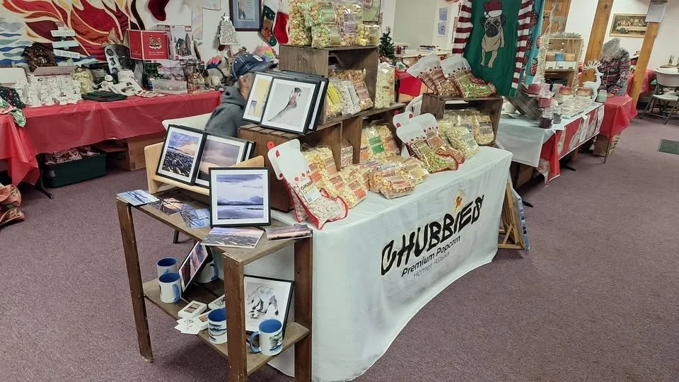 Indoor craft or holiday market with tables displaying framed artwork and basket of packaged popcorn labeled 'Hubbard'.