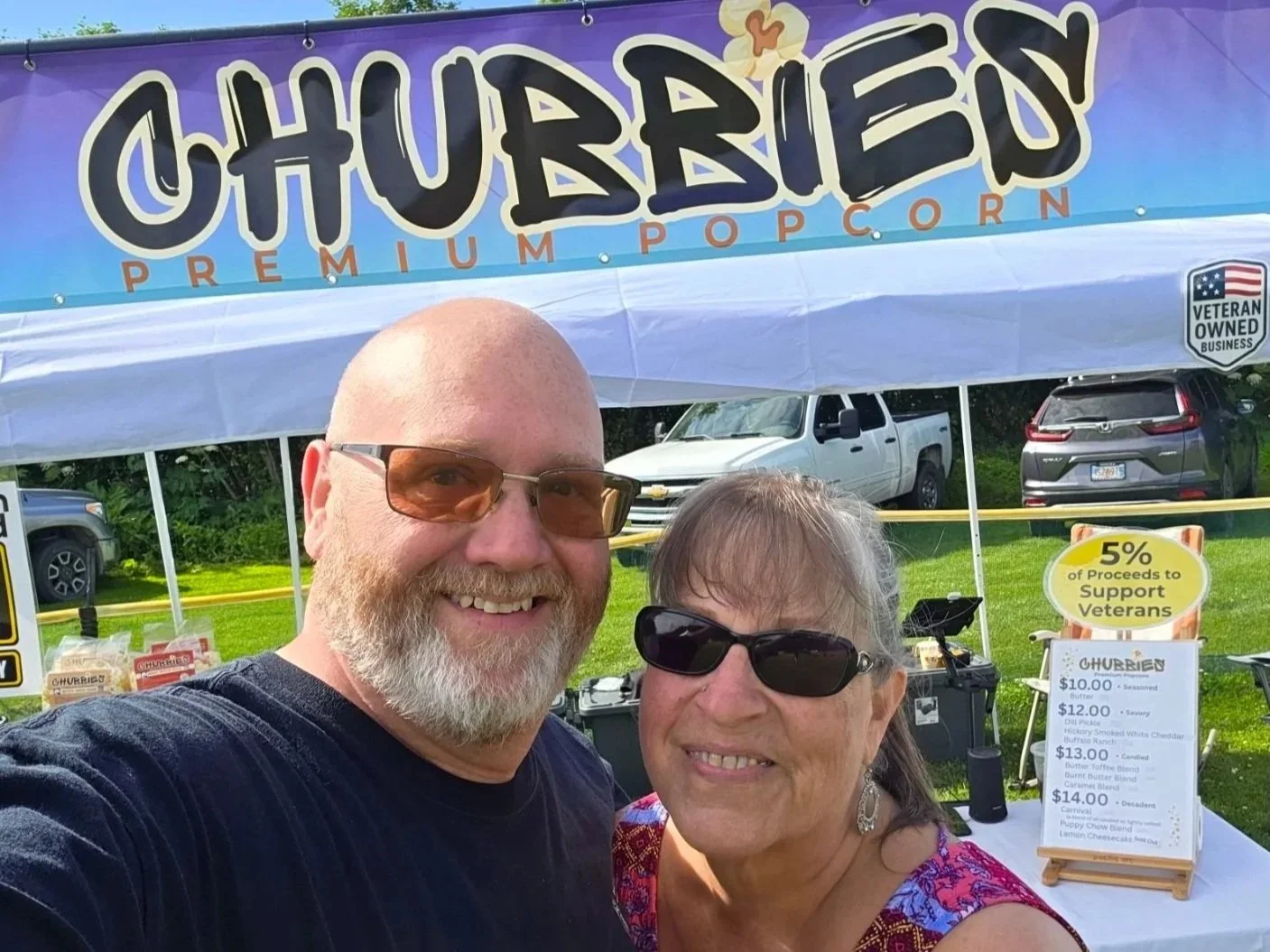A smiling man with a beard wearing glasses and a woman with dark sunglasses and earrings taking a selfie in front of a booth selling gourmet popcorn, under a purple banner with 'CHUBIEU PREMIUM POPCORN' and a sign indicating that 5% of proceeds support veterans.