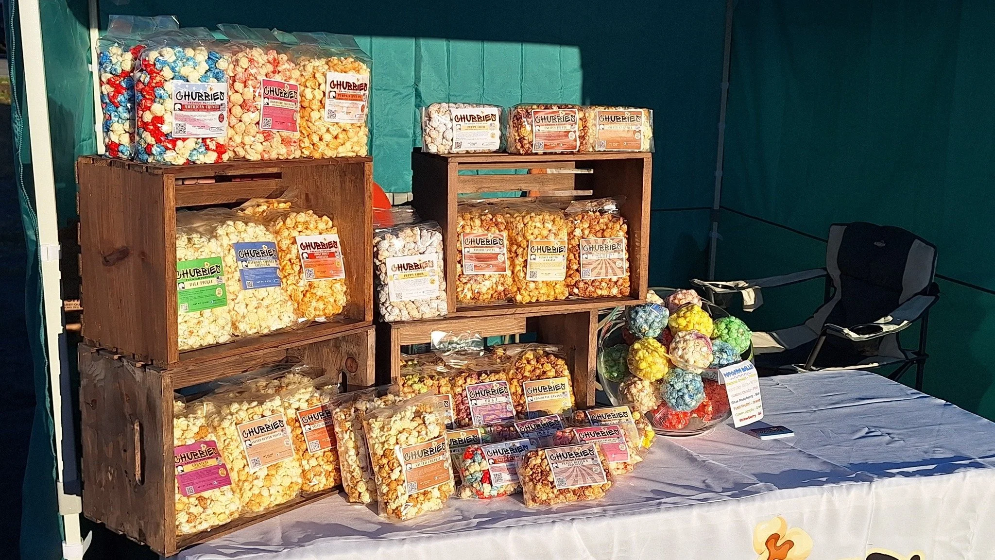 Display of colorful popcorn bags labeled "Chubbie" on wooden crates at a market stall with a bowl of multicolored popcorn balls on a table, and a black folding chair in the background.