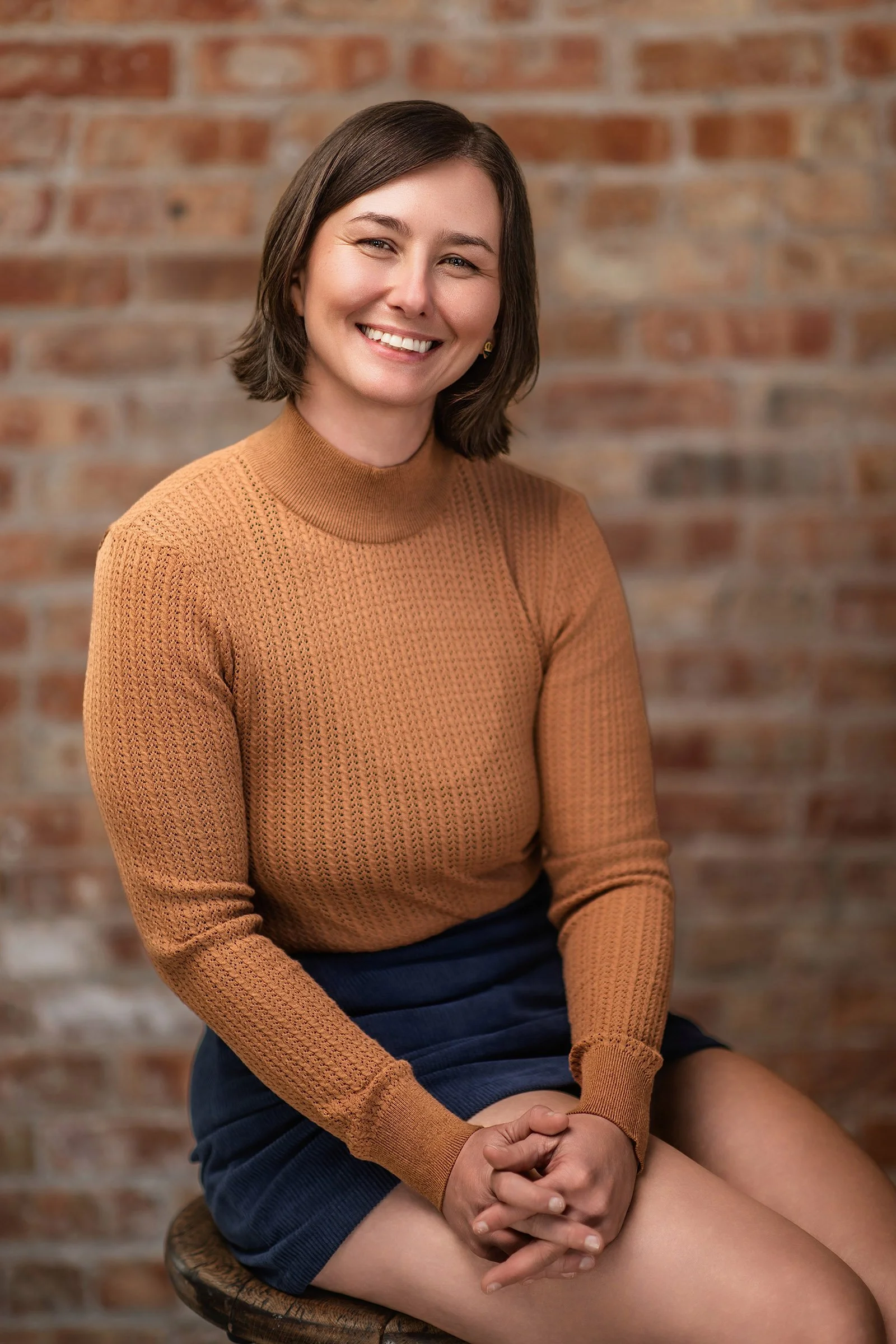 A woman with short brown hair, wearing a brown knitted sweater, sitting on a stool against a brick wall, smiling at the camera.