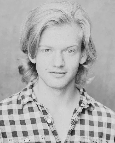 Black and white portrait of a young man with shoulder-length wavy hair, wearing a checkered shirt.