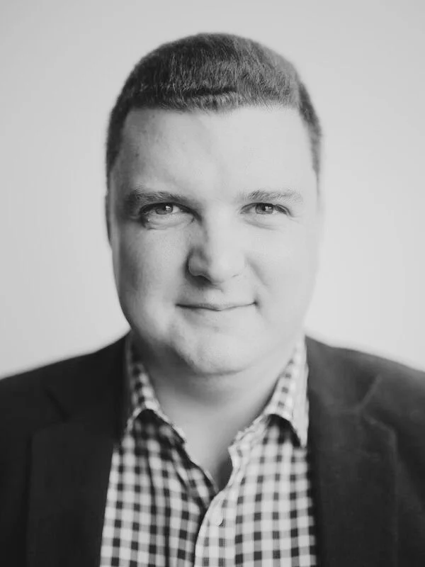 Black and white portrait of a man in business attire with a checked shirt, smiling slightly.