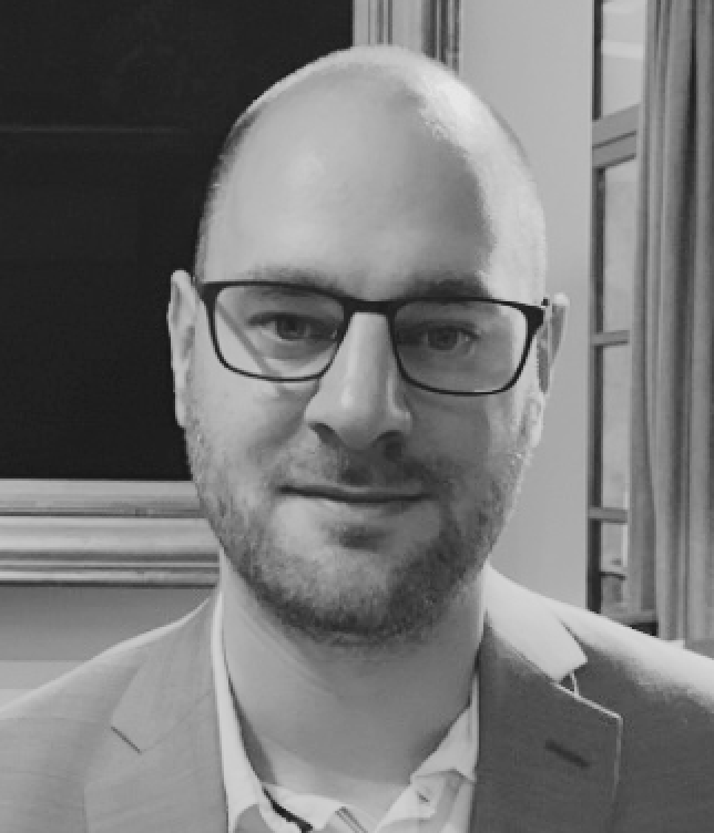 A man with short hair and glasses, wearing a suit jacket and collared shirt, smiling in front of a window with curtains.