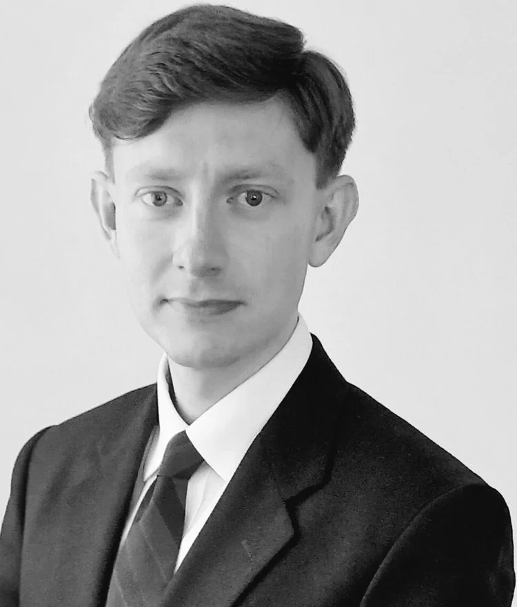 Black and white portrait of a young man with short hair, wearing a suit, tie, and collared shirt, against a plain background.