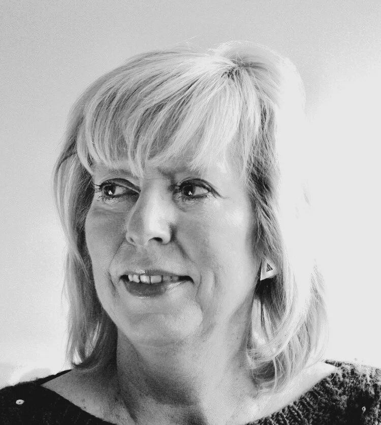 Black-and-white portrait of a middle-aged woman with shoulder-length blonde hair, wearing a dark top with a textured pattern, looking to her left with a slight smile.