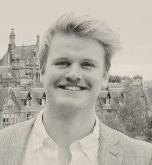 A young man with blonde hair smiling, standing outdoors with buildings and rooftops in the background.