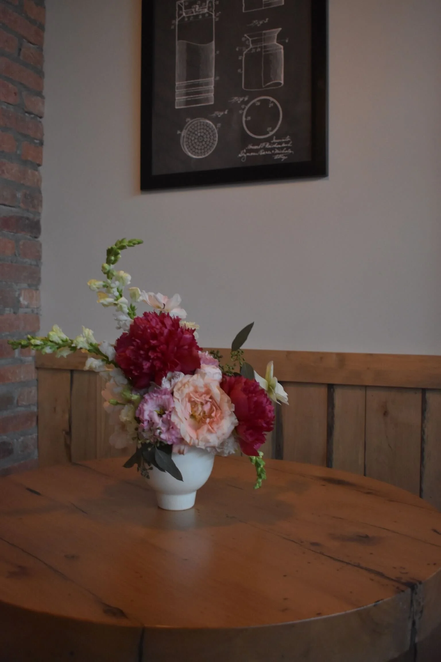 A white vase with pink, red, and white flowers on a wooden table in a room with brick and beige walls, and a framed scientific diagram on the wall.