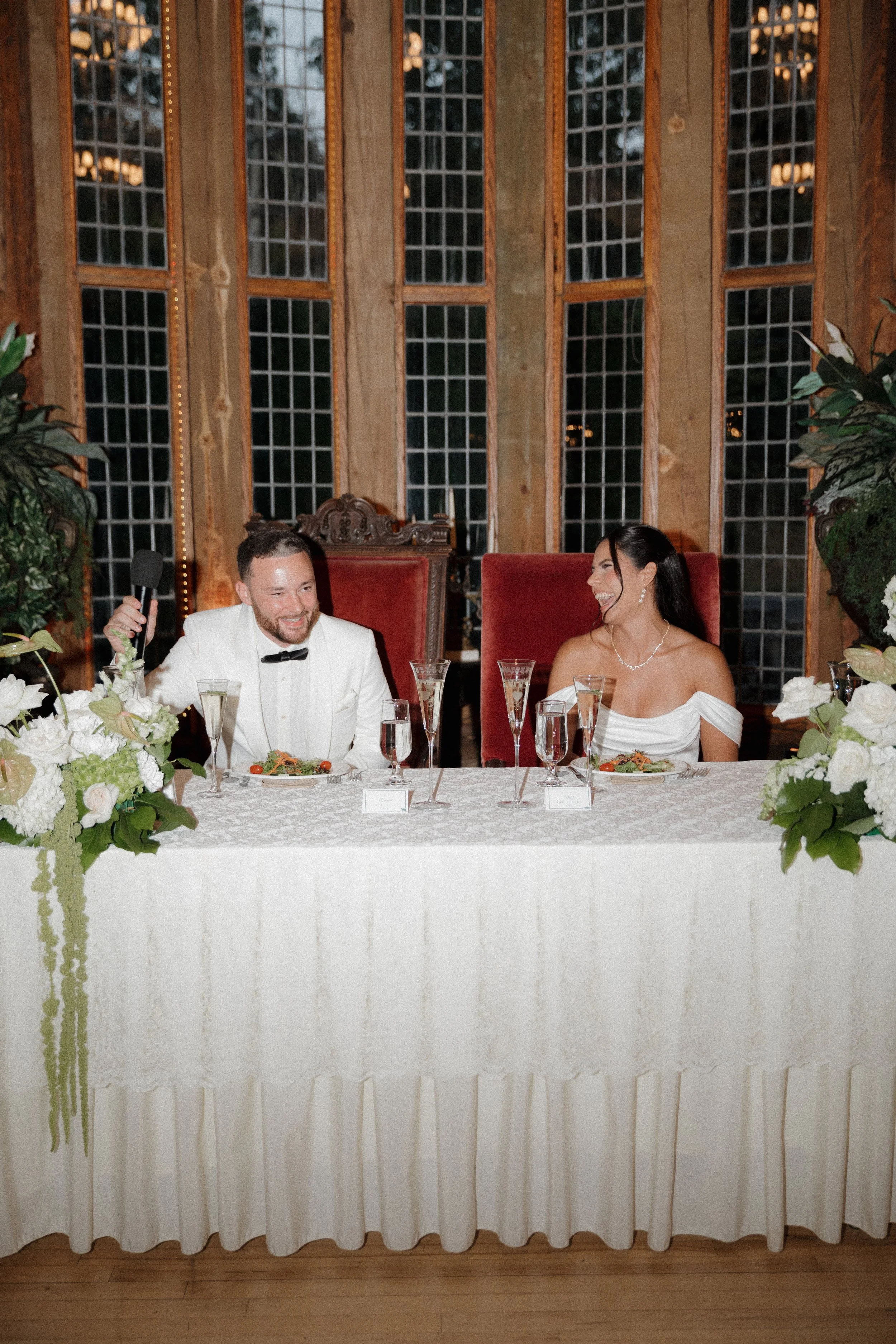 A bride and groom sitting at a wedding reception table, smiling and laughing, with floral arrangements and champagne glasses in front of them.