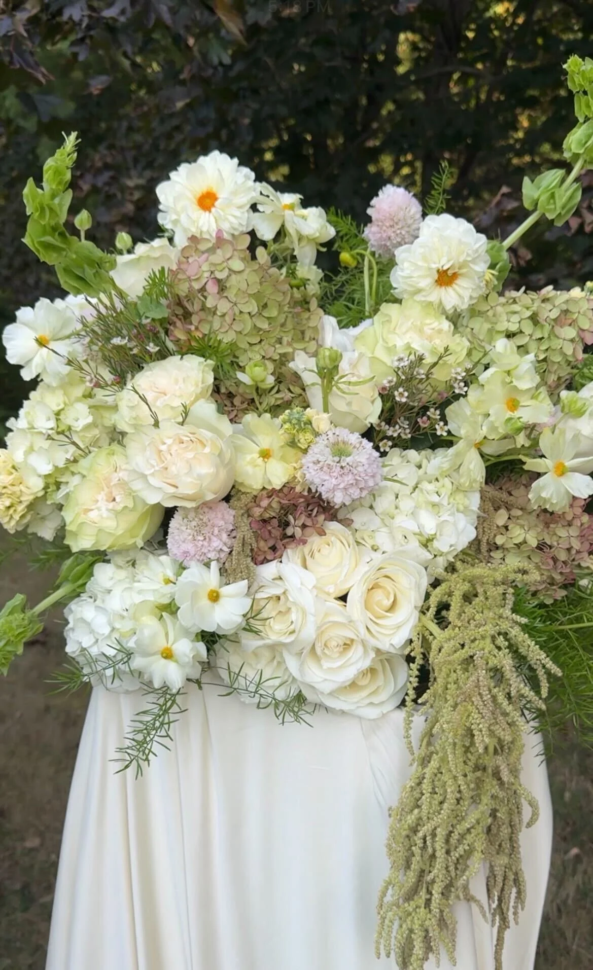 A floral arrangement with white and pale pink flowers in a white vase, set against a dark leafy background.