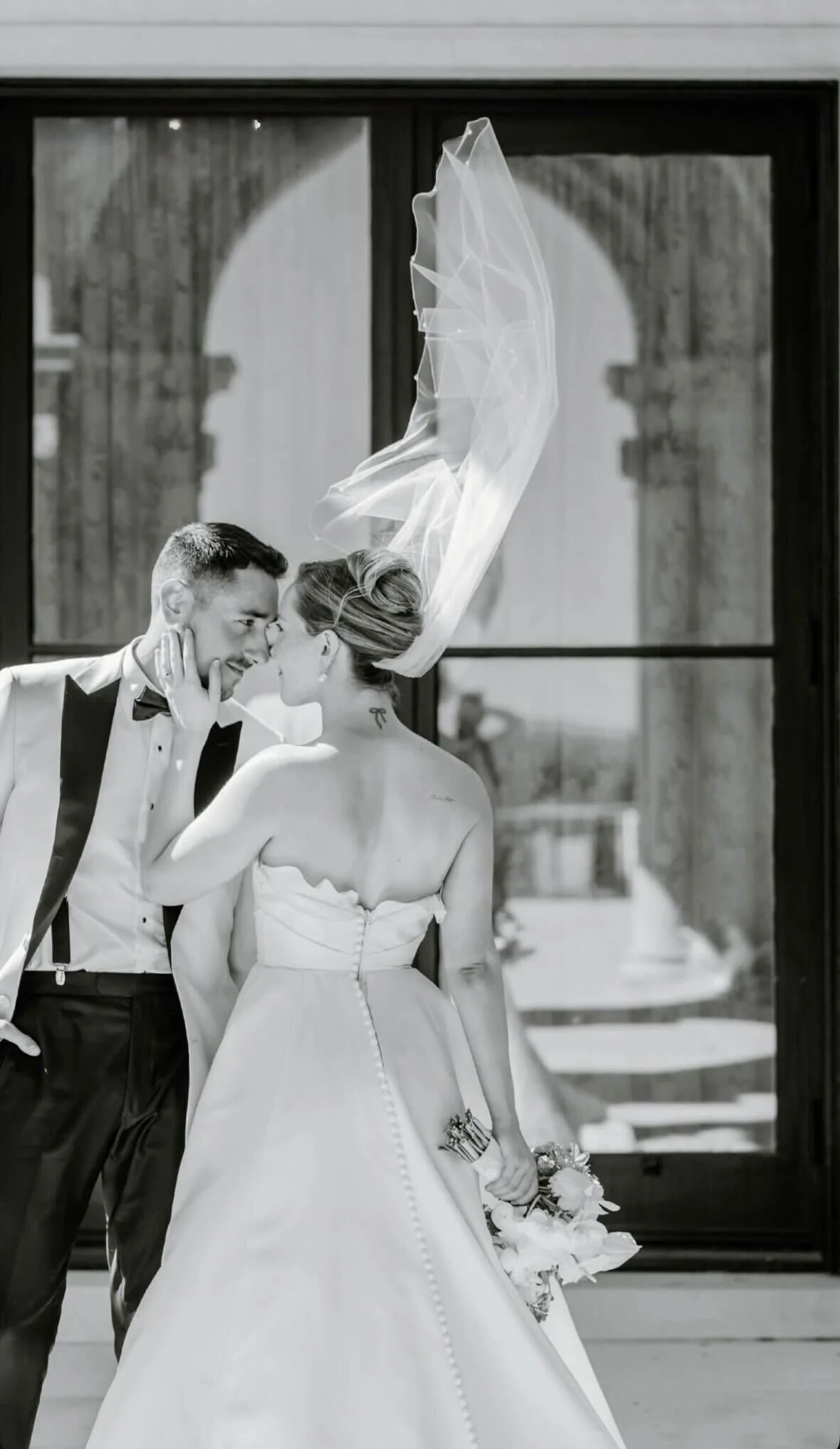 A black and white photograph of a bride and groom in a close, intimate moment. The bride's veil is floating in the air as they lean their foreheads together, with the bride holding a bouquet of flowers.