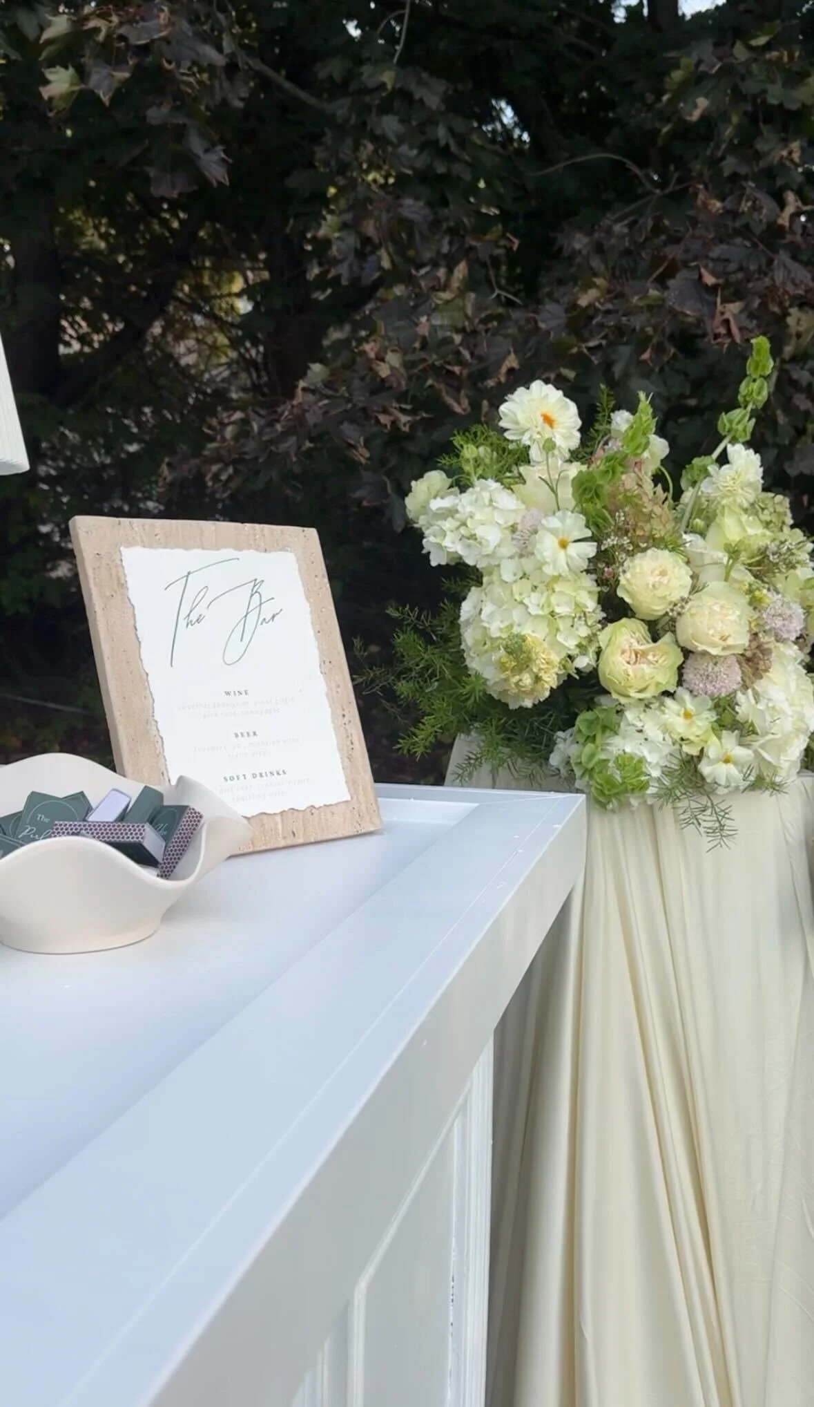 A floral arrangement next to a sign on a white table with a dark leafy background.