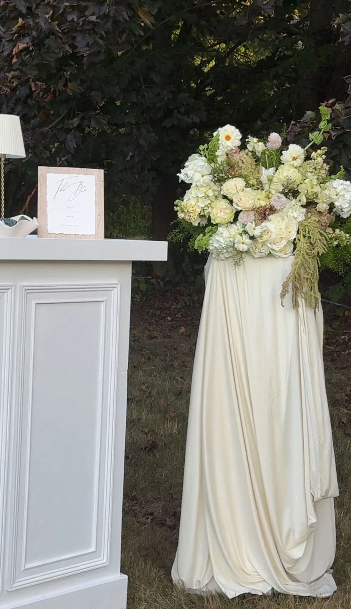 A large floral arrangement of white and pale pink flowers in an off-white draped fabric on a stand outdoors, with a white podium and a framed sign nearby, and dark leafy trees in the background.