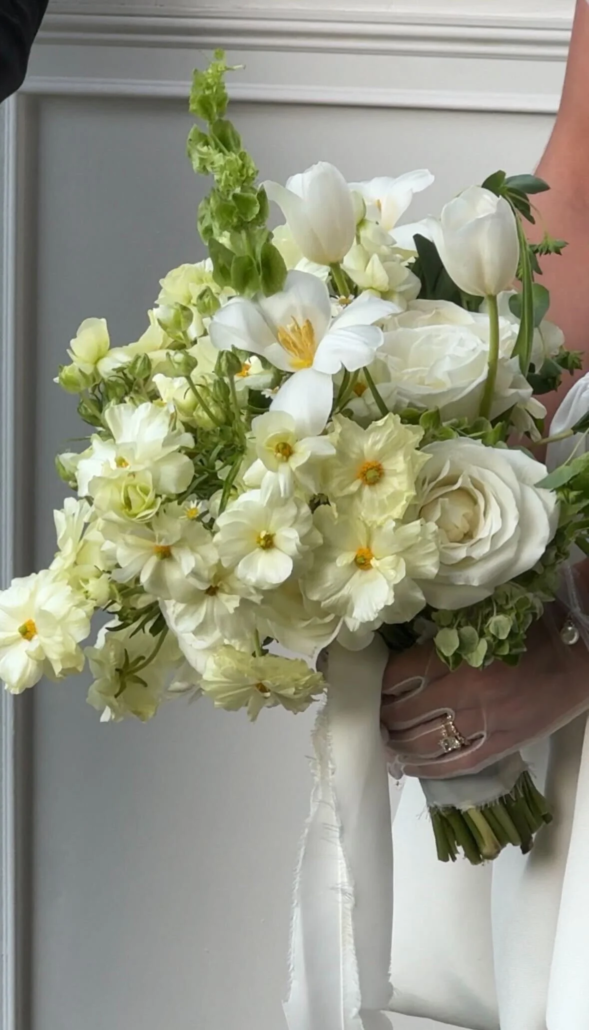A bridal bouquet of white flowers including roses, tulips, and other blooms, wrapped with white ribbon, held by a hand with a ring.