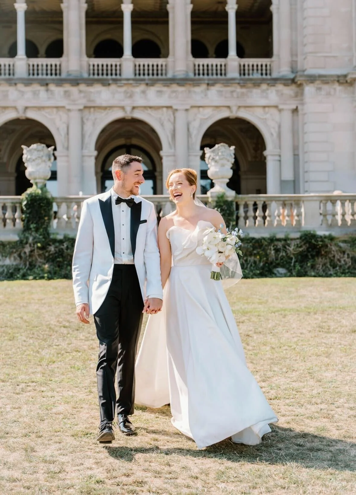 A happy couple in wedding attire walking outdoors in front of a large, ornate building, holding hands and smiling.