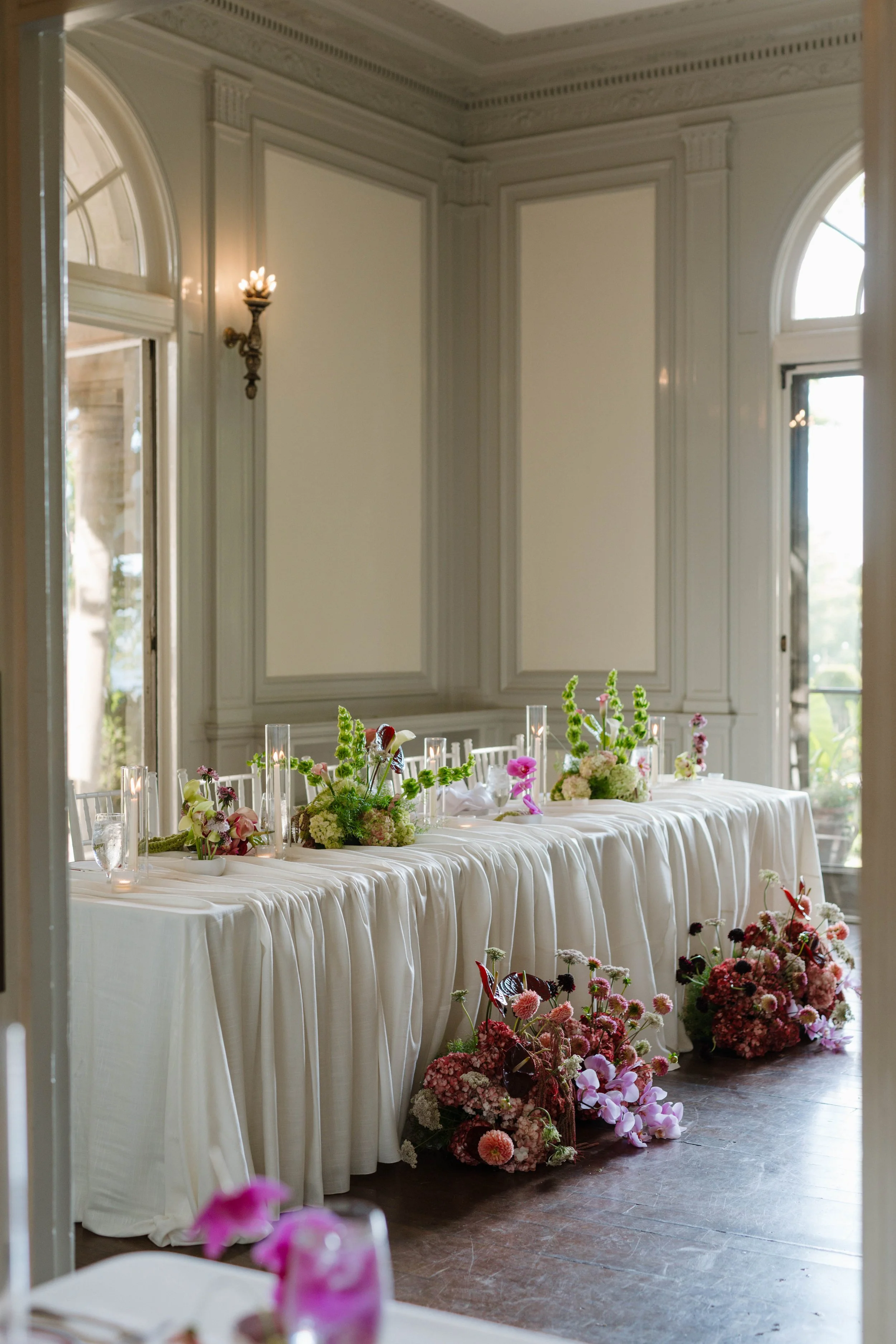 a rectangular table with draping on the top of the table. Ivory tablecloth. Two rectangular centerpieces designed in a green and pink color palette with ivory candles designed throughout. 