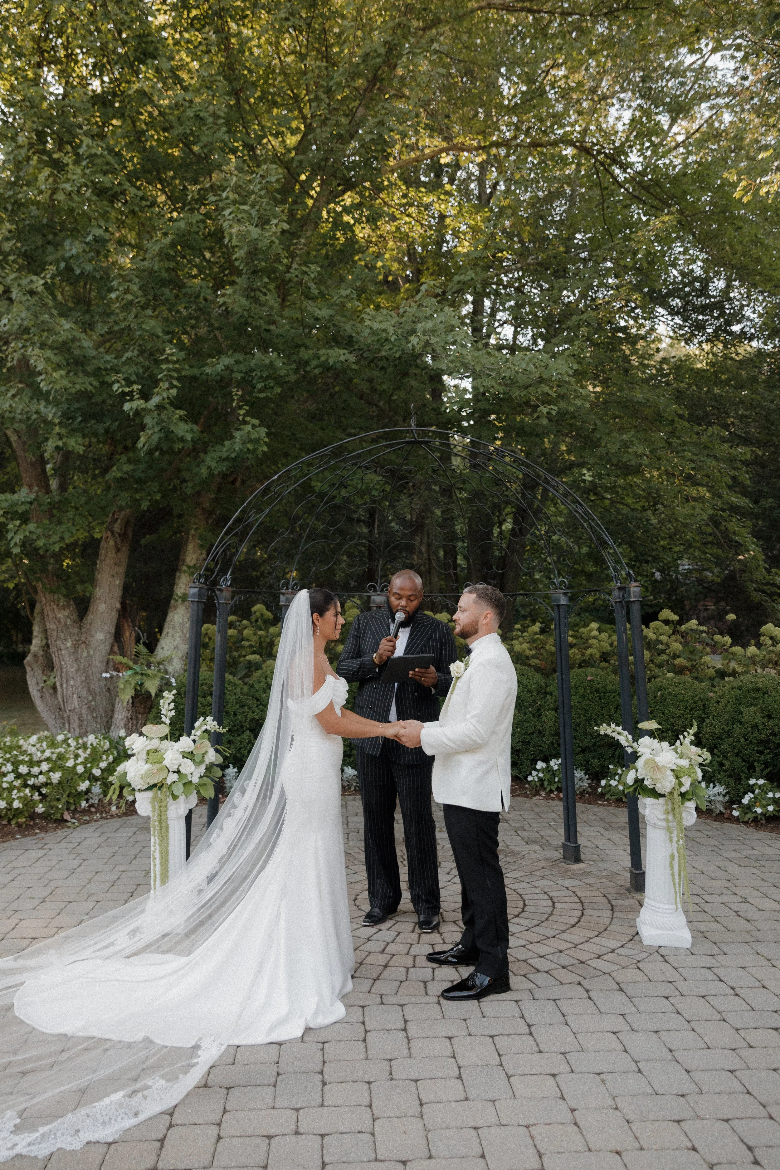 A couple getting married, holding hands, standing under a black metal arch, with an officiant reading from a tablet. The bride wears a white gown with a long train and veil, and the groom wears a white tuxedo jacket. The ceremony is outdoors, surroun