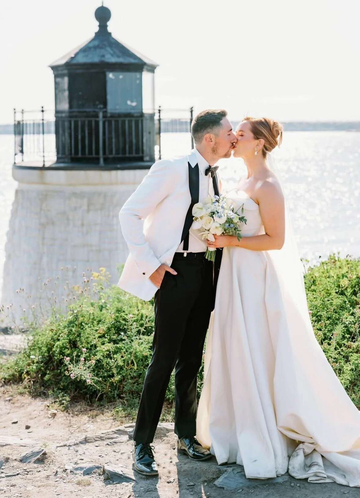 A bride and groom kissing outdoors near a body of water during their wedding, with a lighthouse in the background.