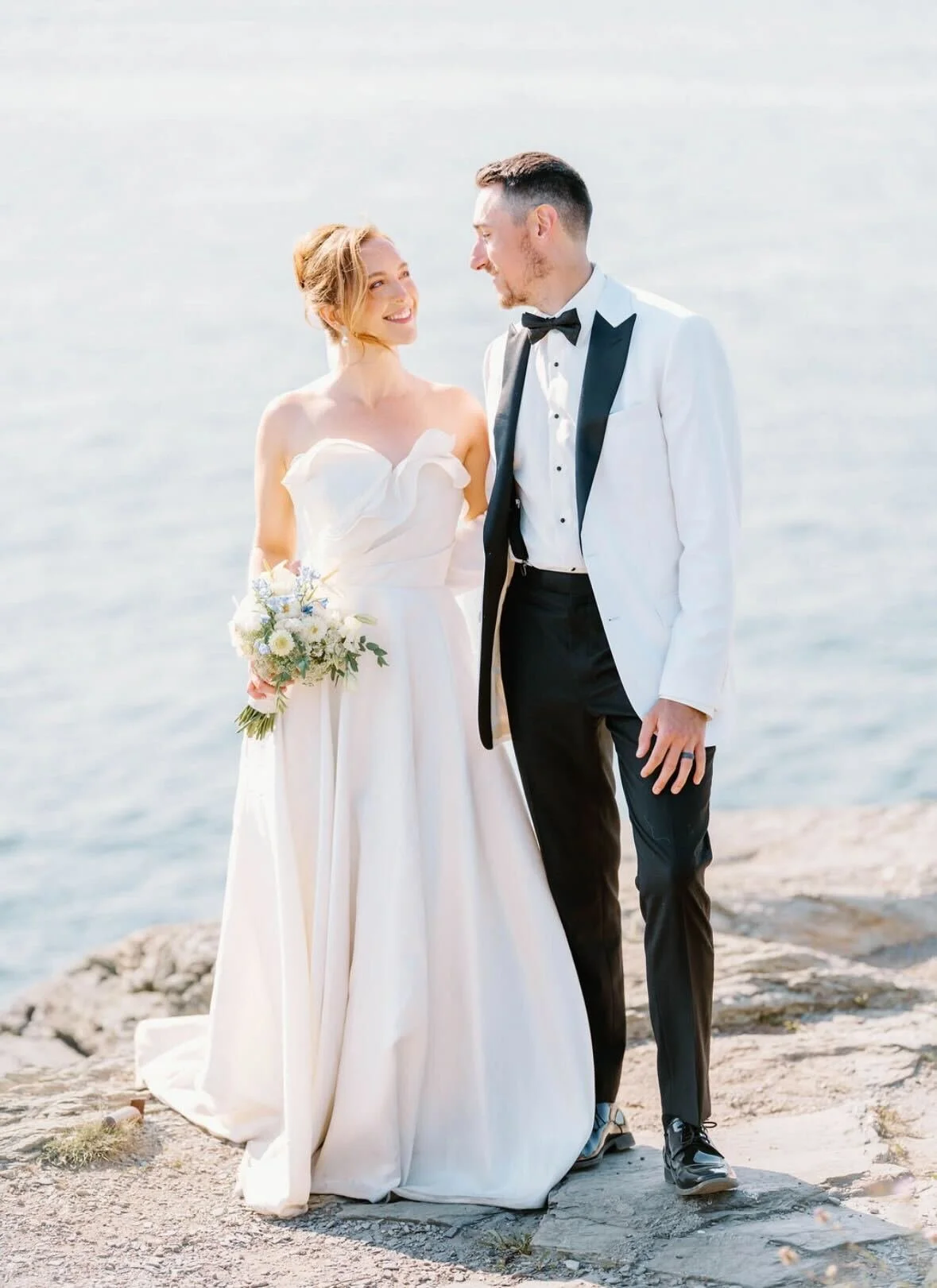 A bride and groom standing on rocks by the water, looking at each other, dressed in wedding attire, smiling.