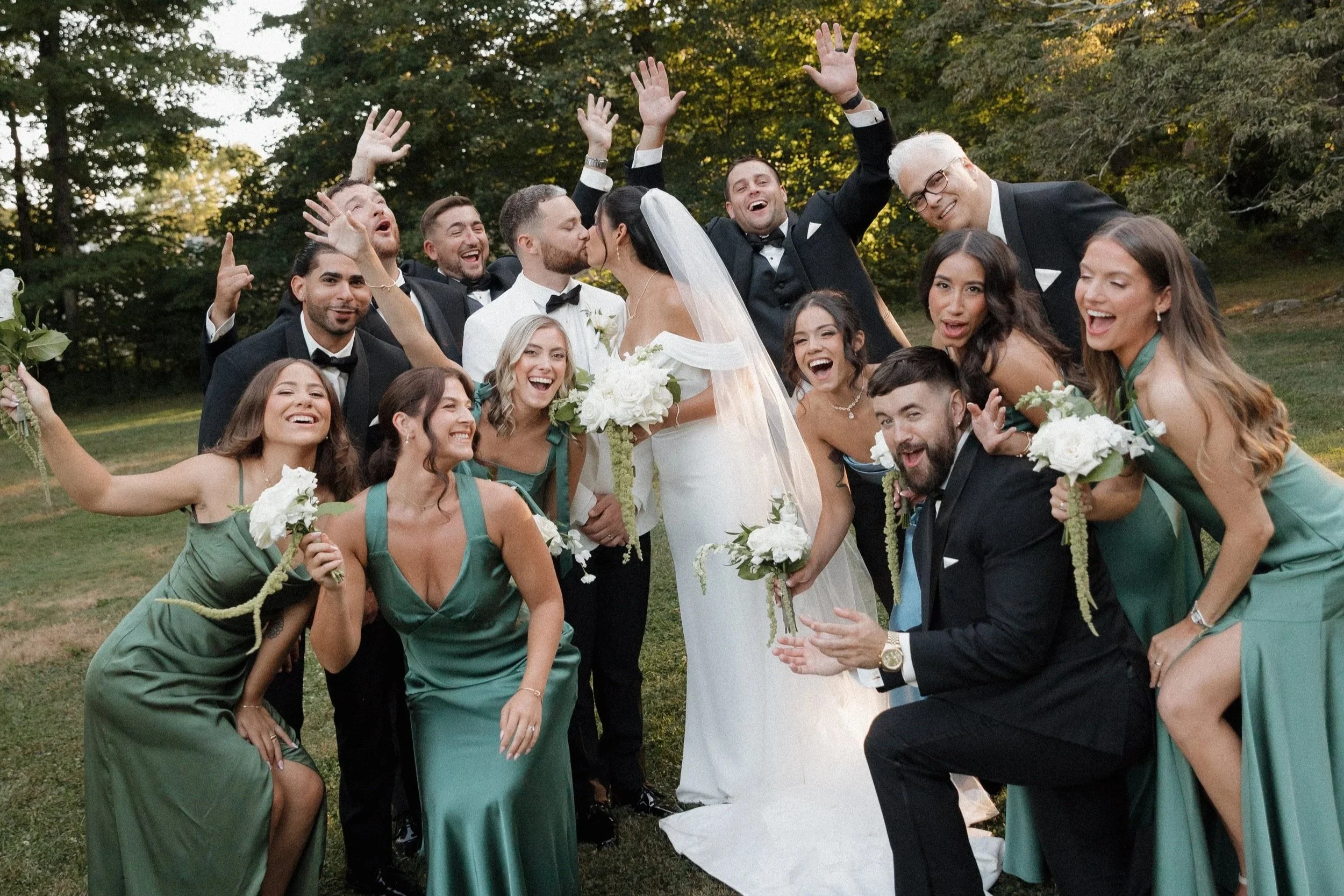 Wedding party with bride and groom kissing, surrounded by bridesmaids in teal dresses and groomsmen in black tuxedos, outdoors in a park with trees in the background.