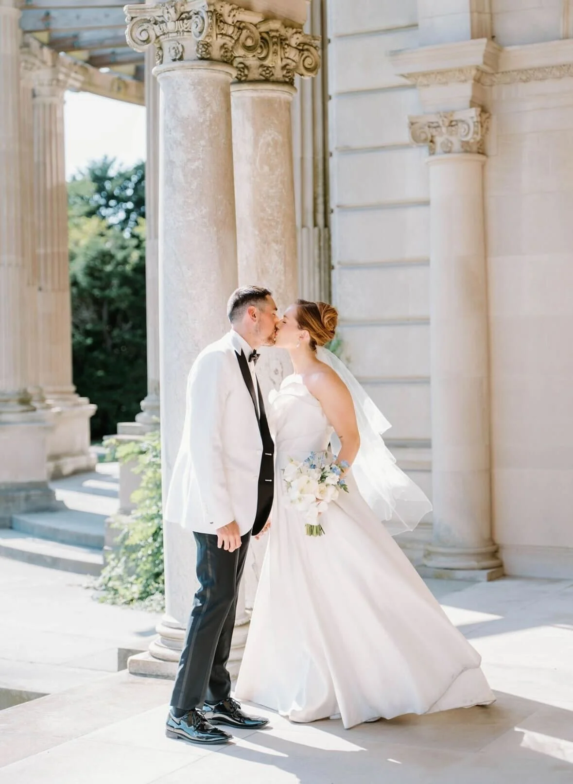 A bride and groom sharing a kiss outdoors in front of classical columns at a wedding.