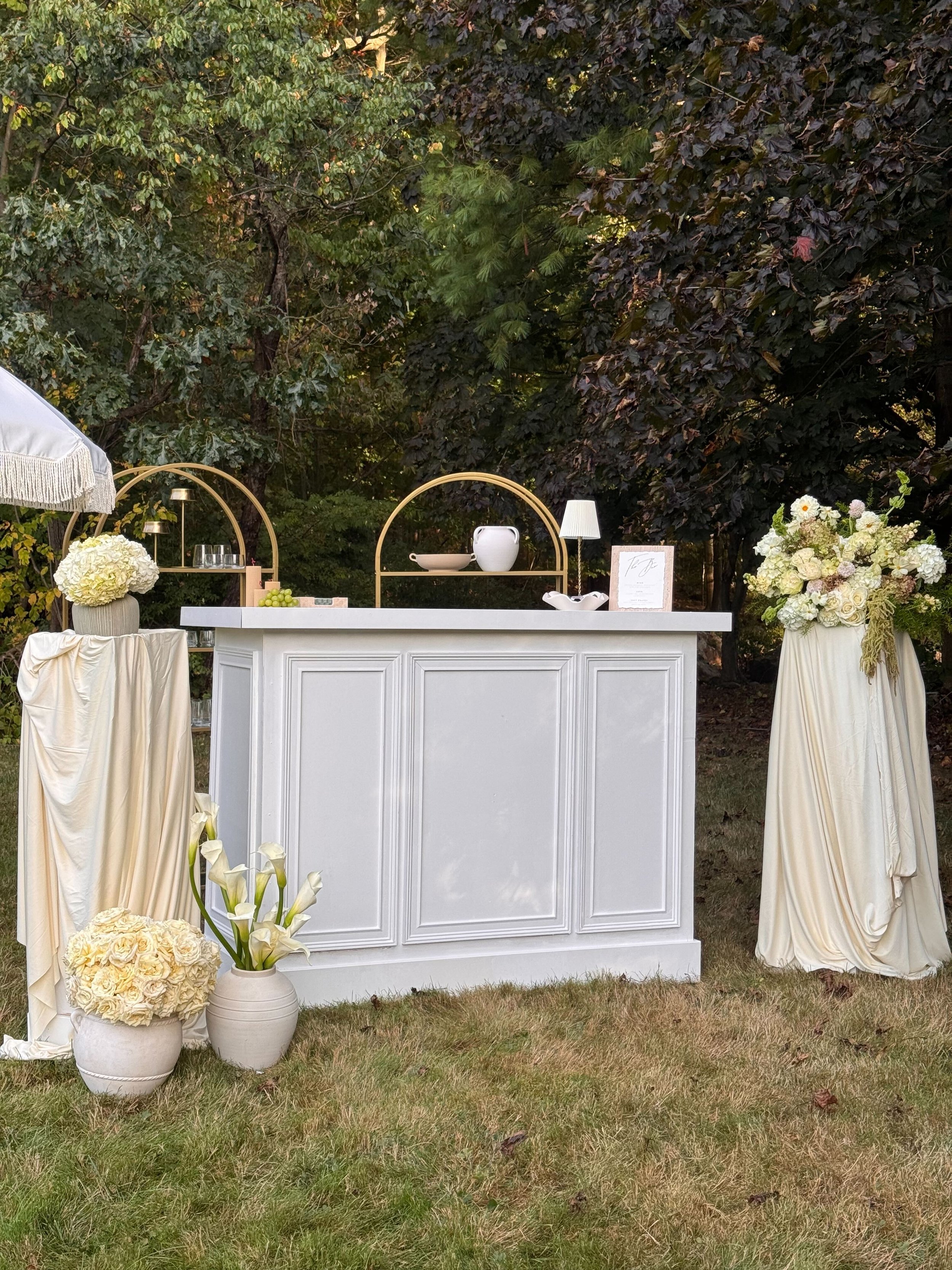Outdoor bar setup with a white counter and floral arrangements in white vases, a small lamp, and decorative elements, surrounded by trees and foliage.
