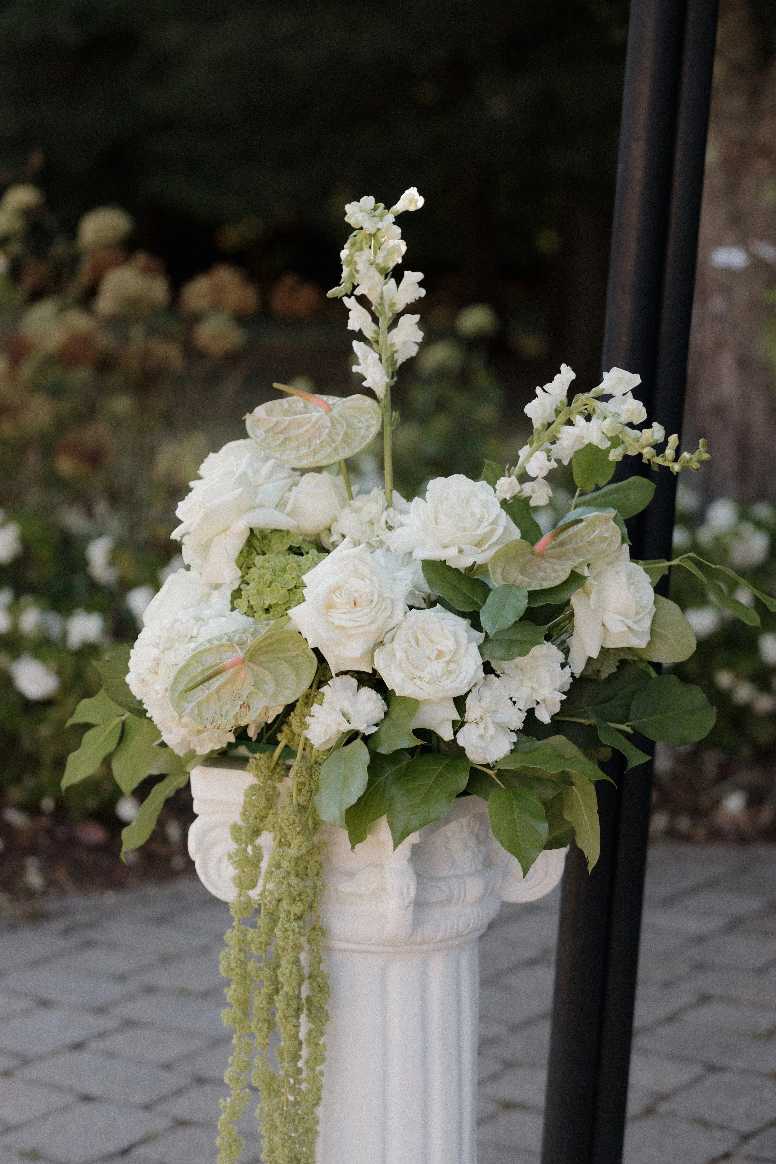 A white floral arrangement in a white vase with roses, anthuriums, and greenery, placed on a white pedestal outside.