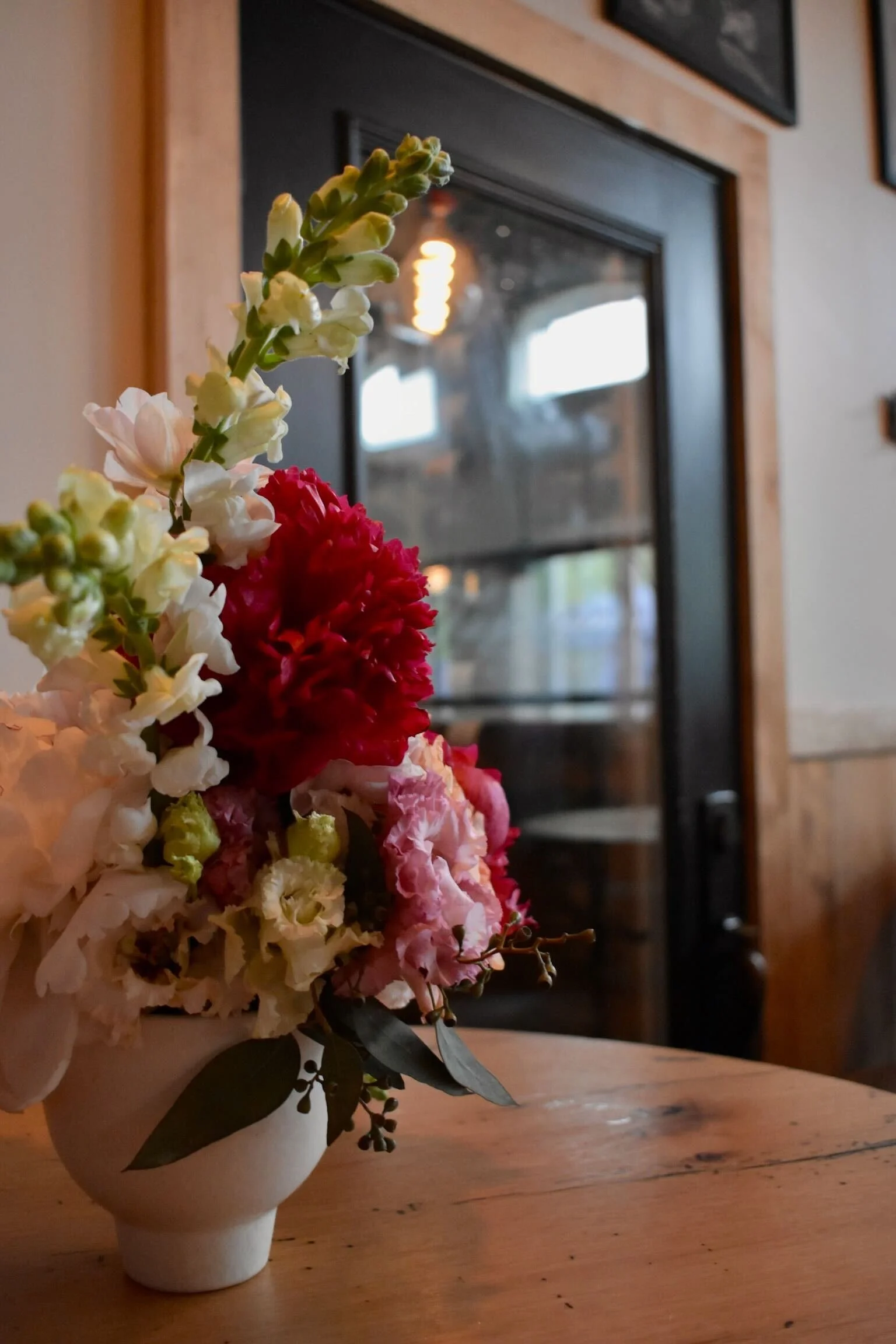 A white ceramic vase with a floral arrangement, including white, pink, and red flowers, sits on a wooden table in a cozy indoor setting. A black-framed glass door is in the background.