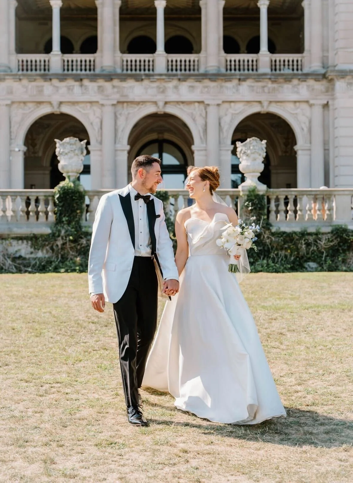 Bride and groom walking hand in hand outdoors in front of a historic building, smiling at each other, with the bride holding a bouquet of white flowers.