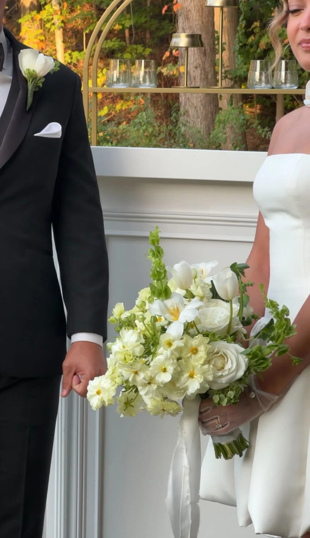 Close-up of a bride holding a bouquet of white flowers at a wedding ceremony, with a groom in a black tuxedo standing nearby.