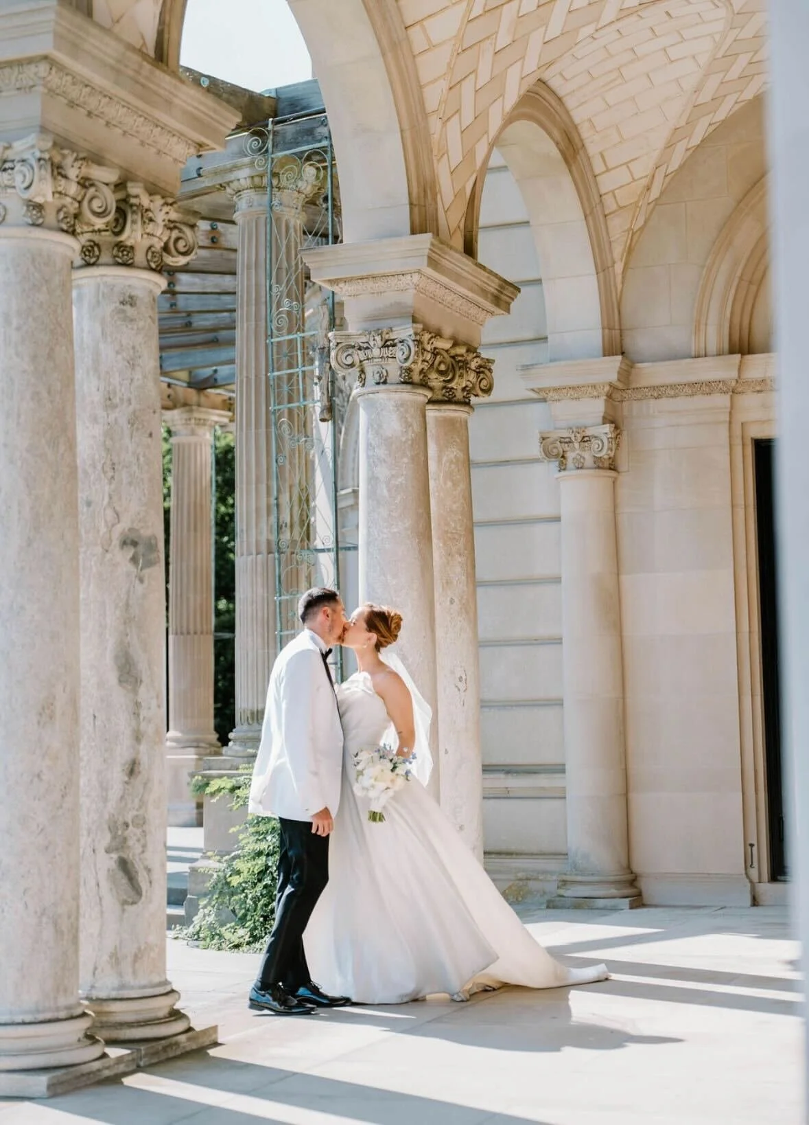A bride and groom sharing a kiss outdoors beneath classical columns and arches, with sunlight casting shadows.
