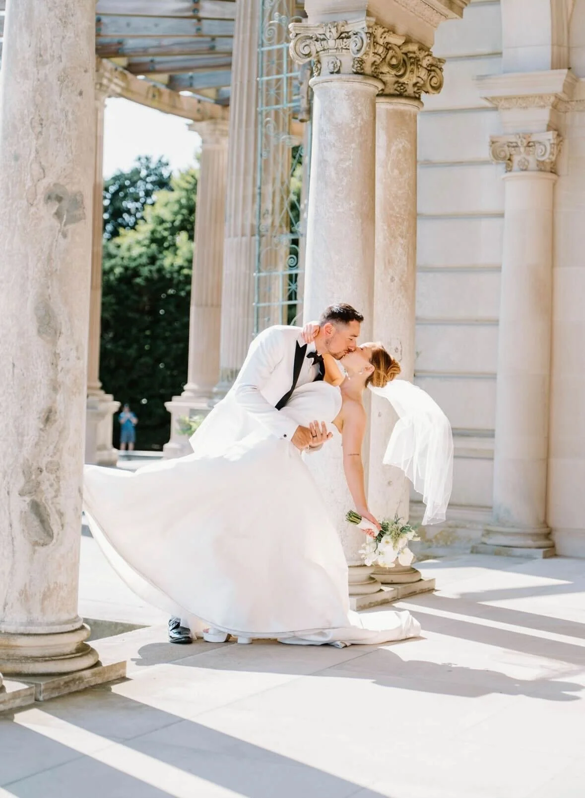 A bride and groom kissing outside among classical columns and marble stairs, with the groom holding the bride in a dip.