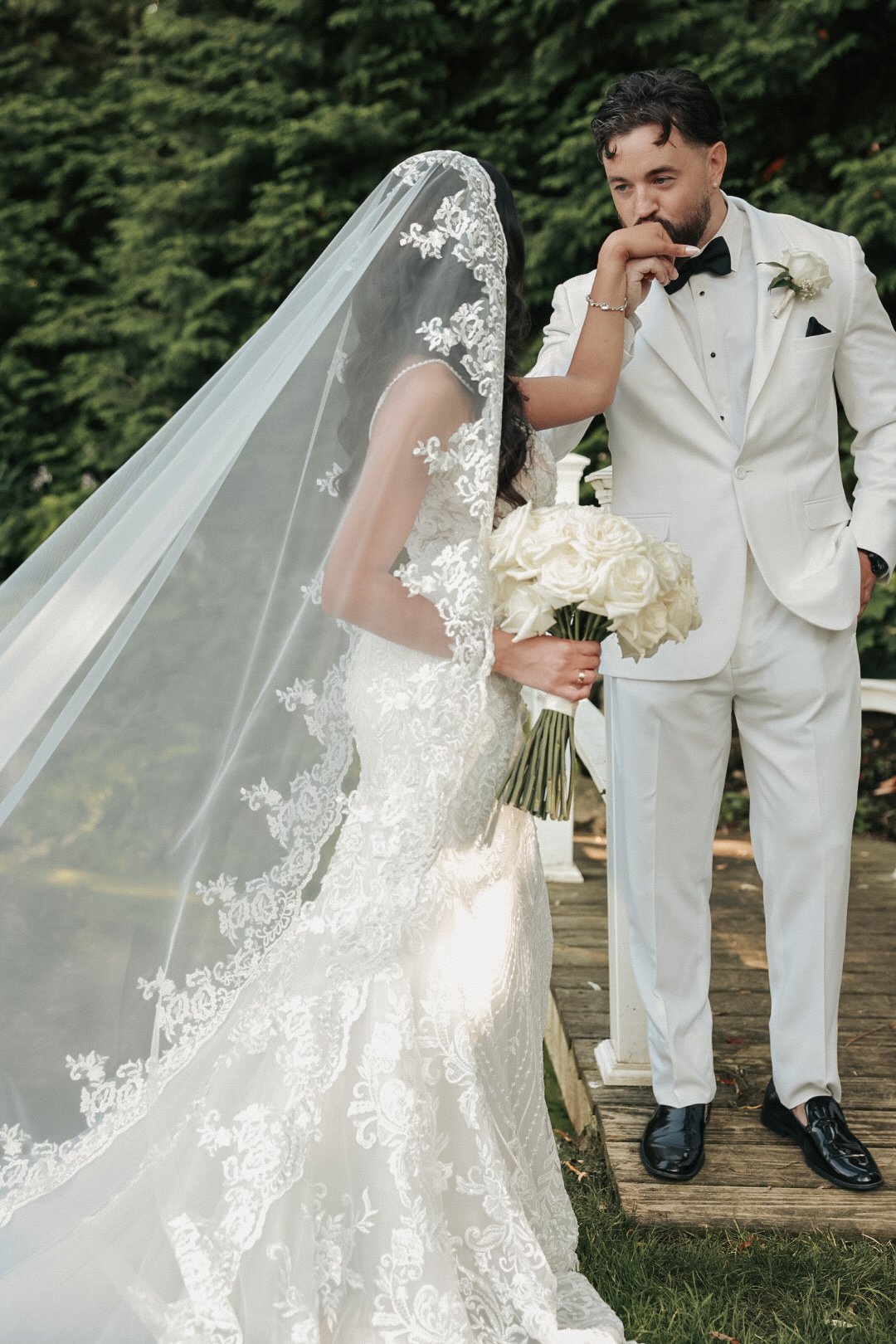 couple dressed in a white tux and a white bridal gown. Bride is holding a playa blanca rose bridal bouquet and the husband is kissing her hand. 