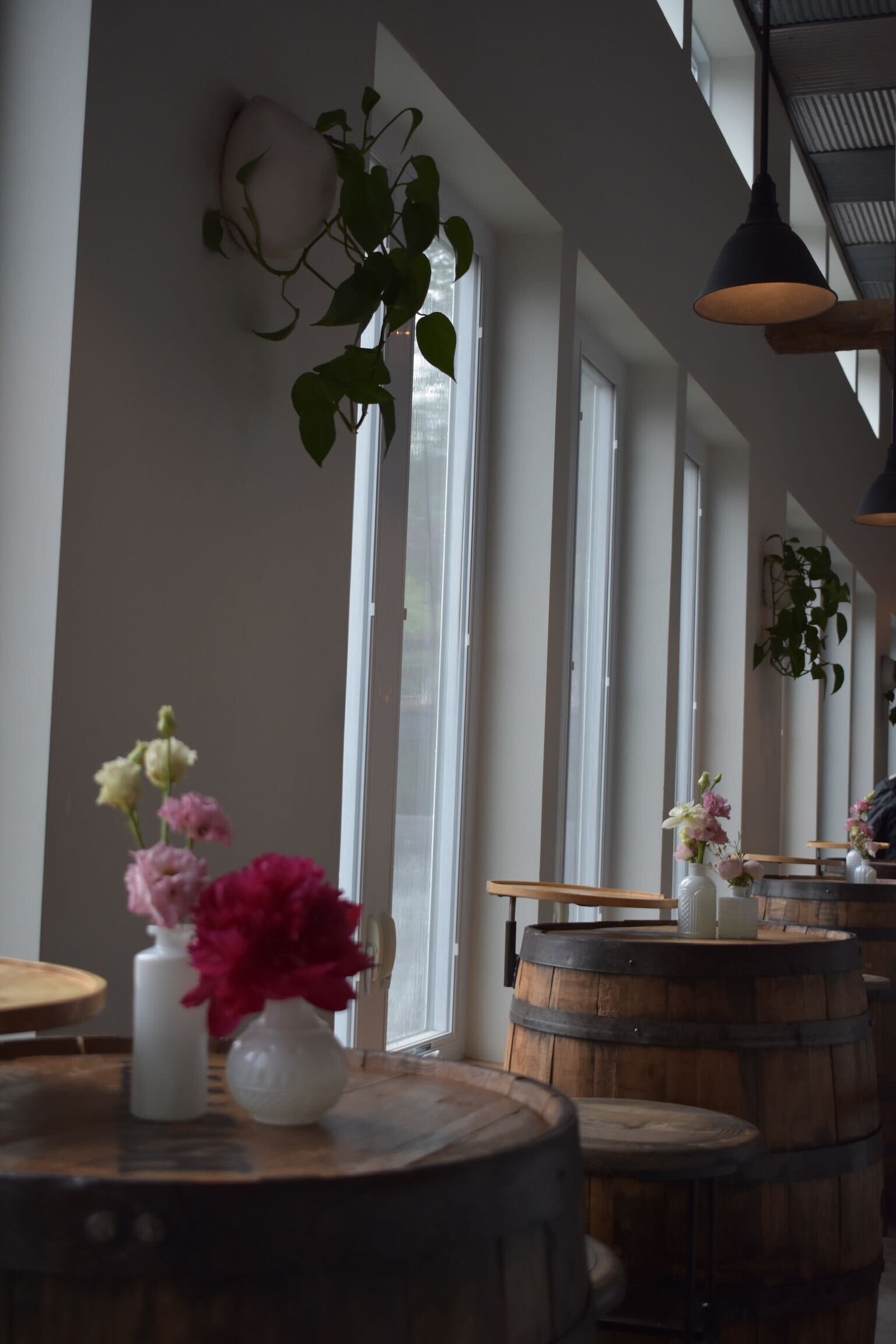 Interior of a modern cafe or restaurant with wooden barrel tables, each decorated with small vases of pink and white flowers, and large windows allowing natural light to enter.