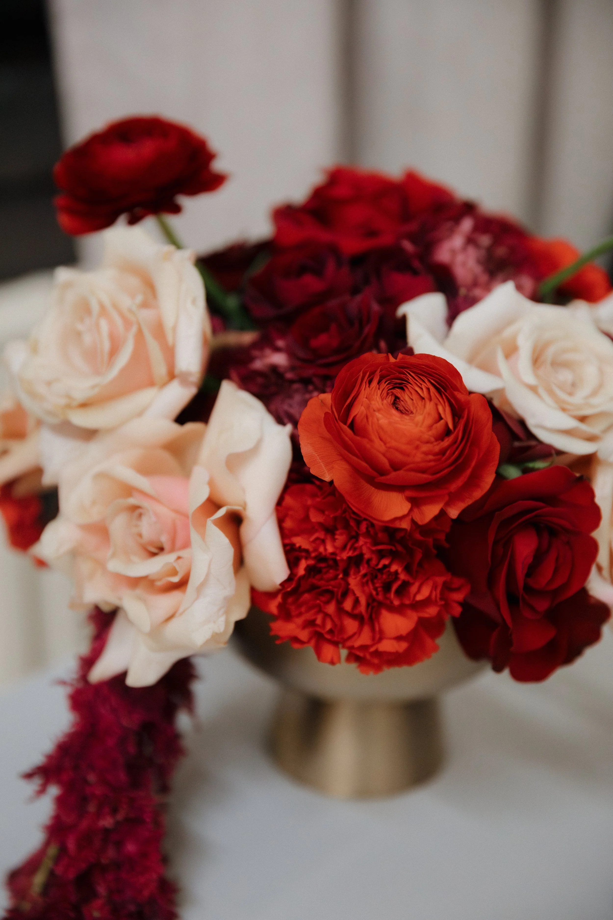 Compote vase arrangement in a gold compote. Designed with red roses, red ranunculus, soft blush pink roses, red spray roses, and red carnations. Draped with red amaranths. 