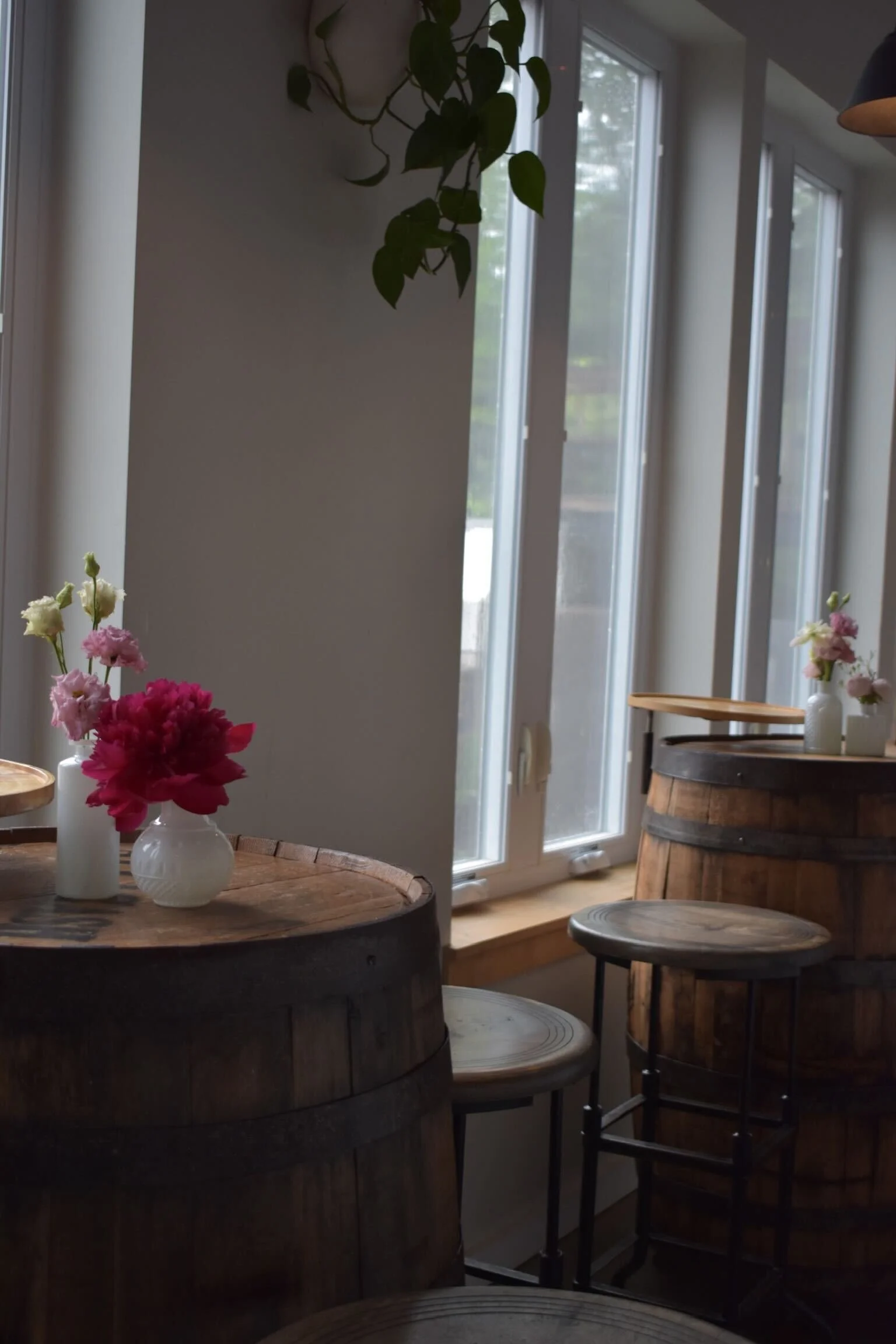 Interior of a cafe with wooden barrel tables, stools, and vases with pink and white flowers near large windows.