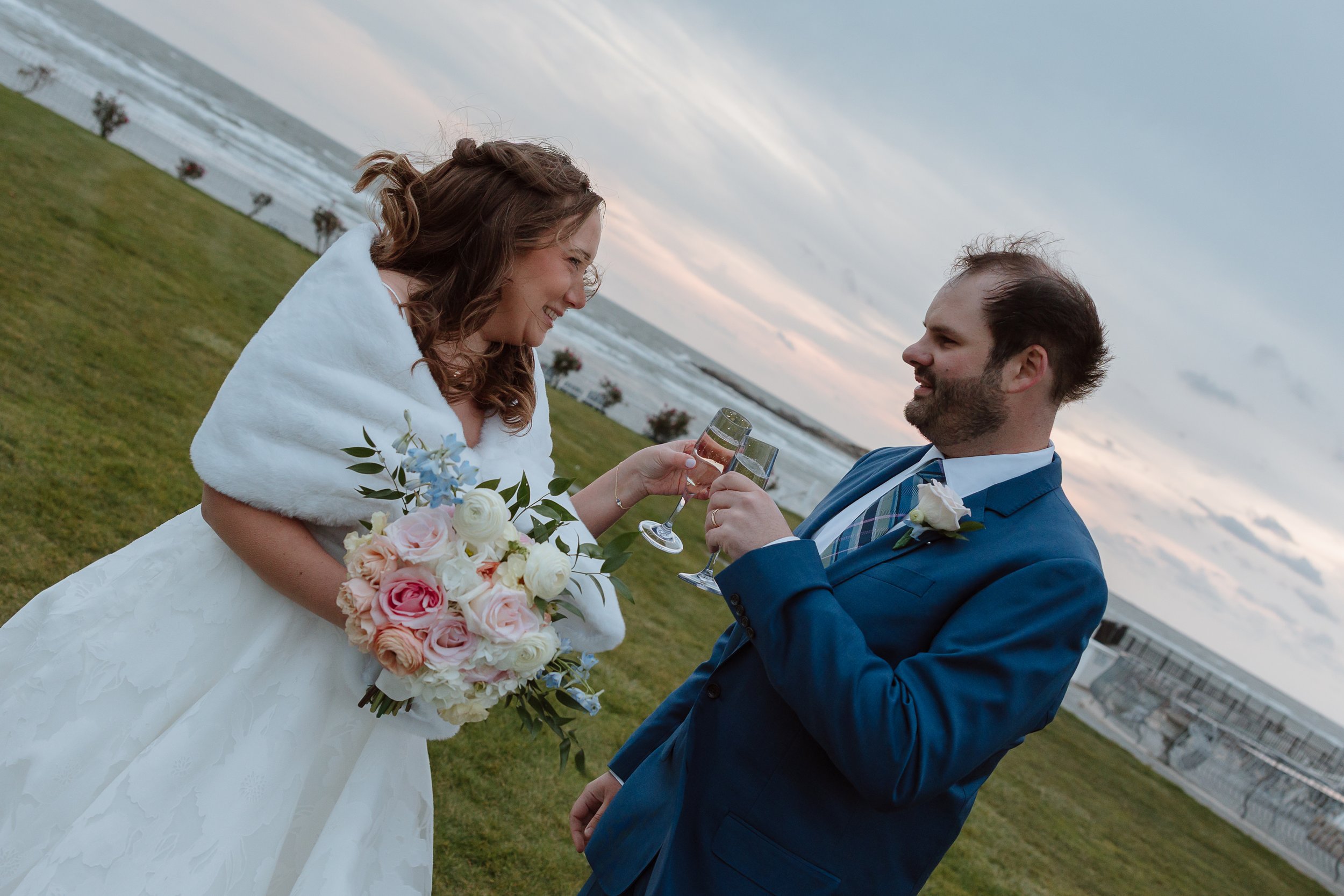 Bride and groom cheer-sing, holding a garden style bridal bouquet. Bride is wearing a white fur overlay coat and the Groom is in a navy blue tux with a soft blush rose boutonnière.