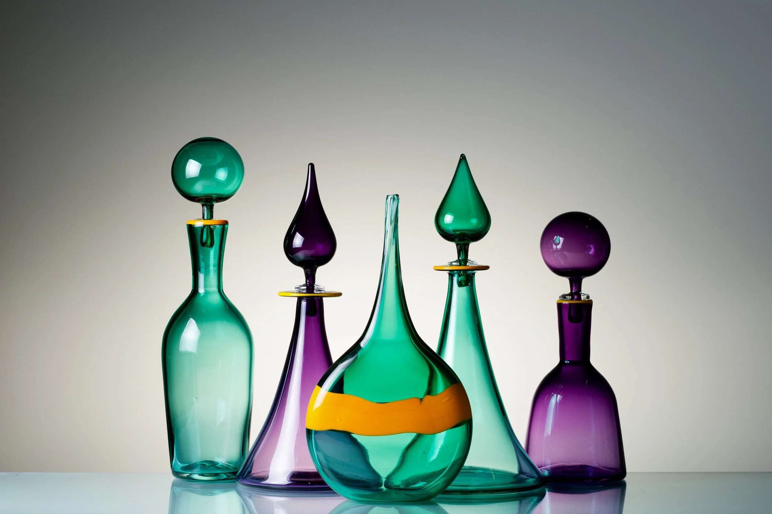 Colorful glass vases with various shapes and colors, including green, purple, and yellow, arranged on a reflective surface against a gradient background.