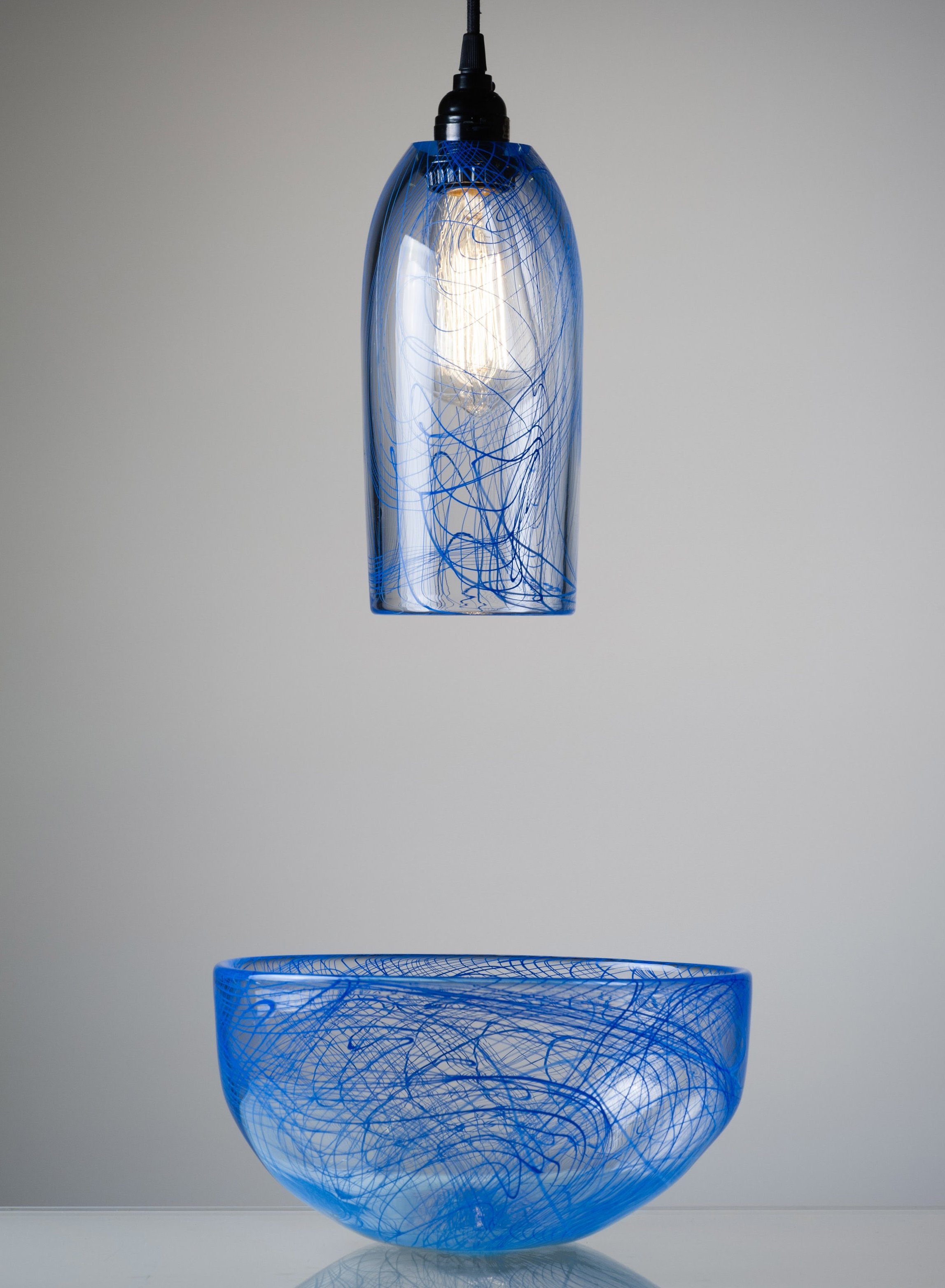 A hanging glass pendant light with blue swirls and lines, above a matching blue glass bowl with similar swirling patterns, set against a plain light-colored background.
