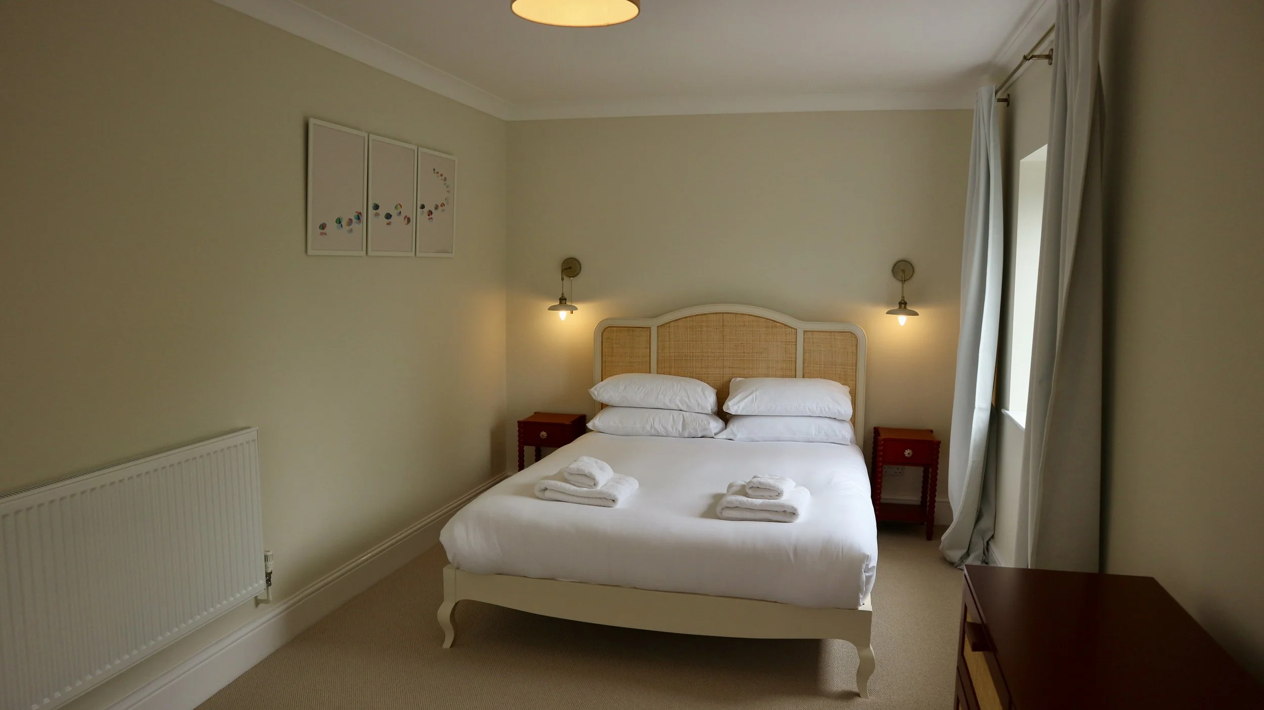 A neatly made bed with white linens, four pillows, and folded towels on top, in a softly lit bedroom with beige walls, two small nightstands, wall-mounted lights, a window with white curtains, and a piece of wall art. 