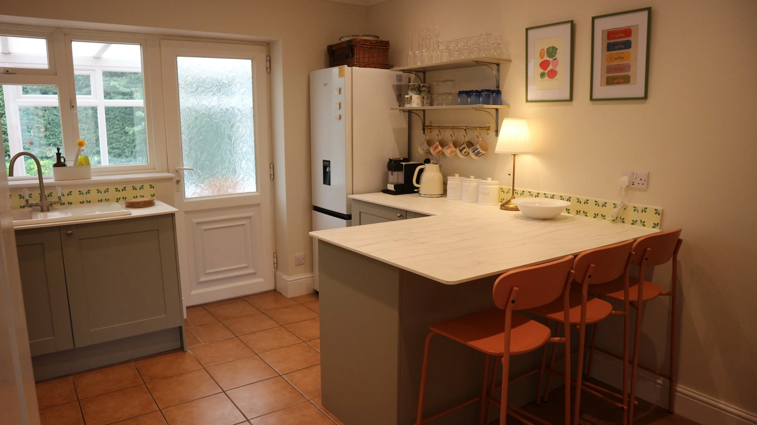 A  kitchen with a white door leading outside, a small grey cabinet under a sink, a window above the sink, and a white marble countertop with three orange barstools. The countertop has a bowl, a lamp, and kitchen appliances, with framed artwork and gl