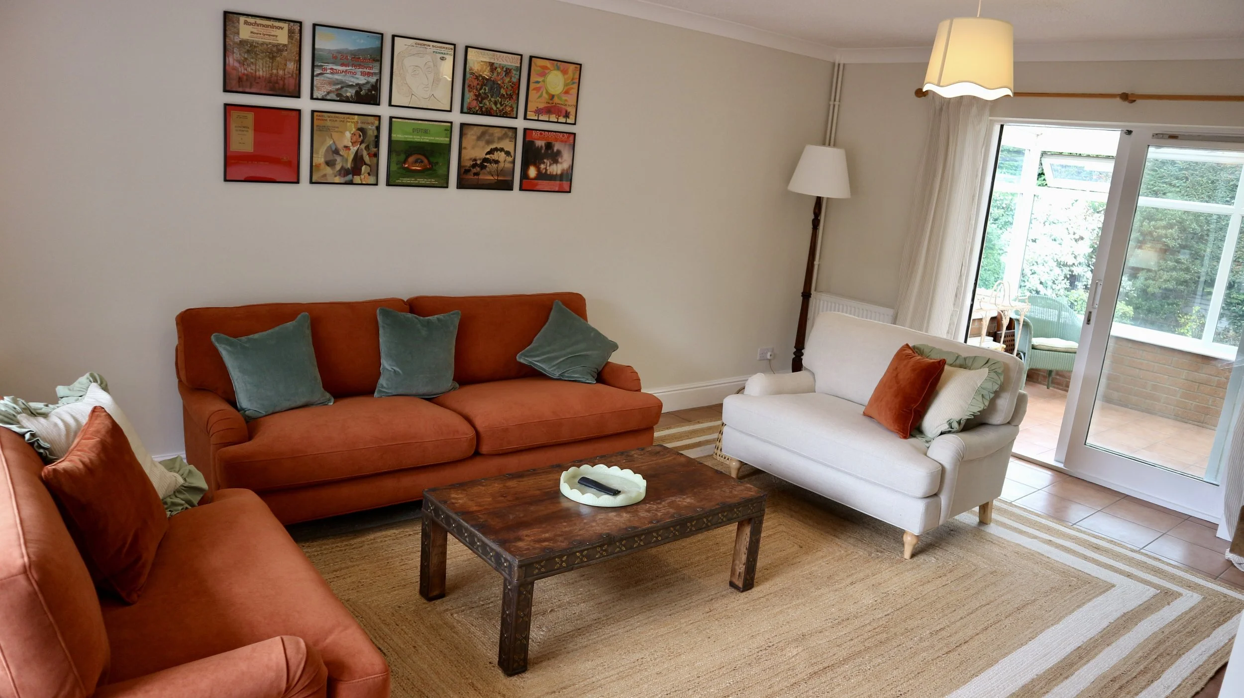 Living room with two orange sofas, a white armchair, a wooden coffee table, framed pictures on the wall, a large window with curtains, and a glass door leading to the conservatory diner. Blackbird Cottage, recently refurbished.