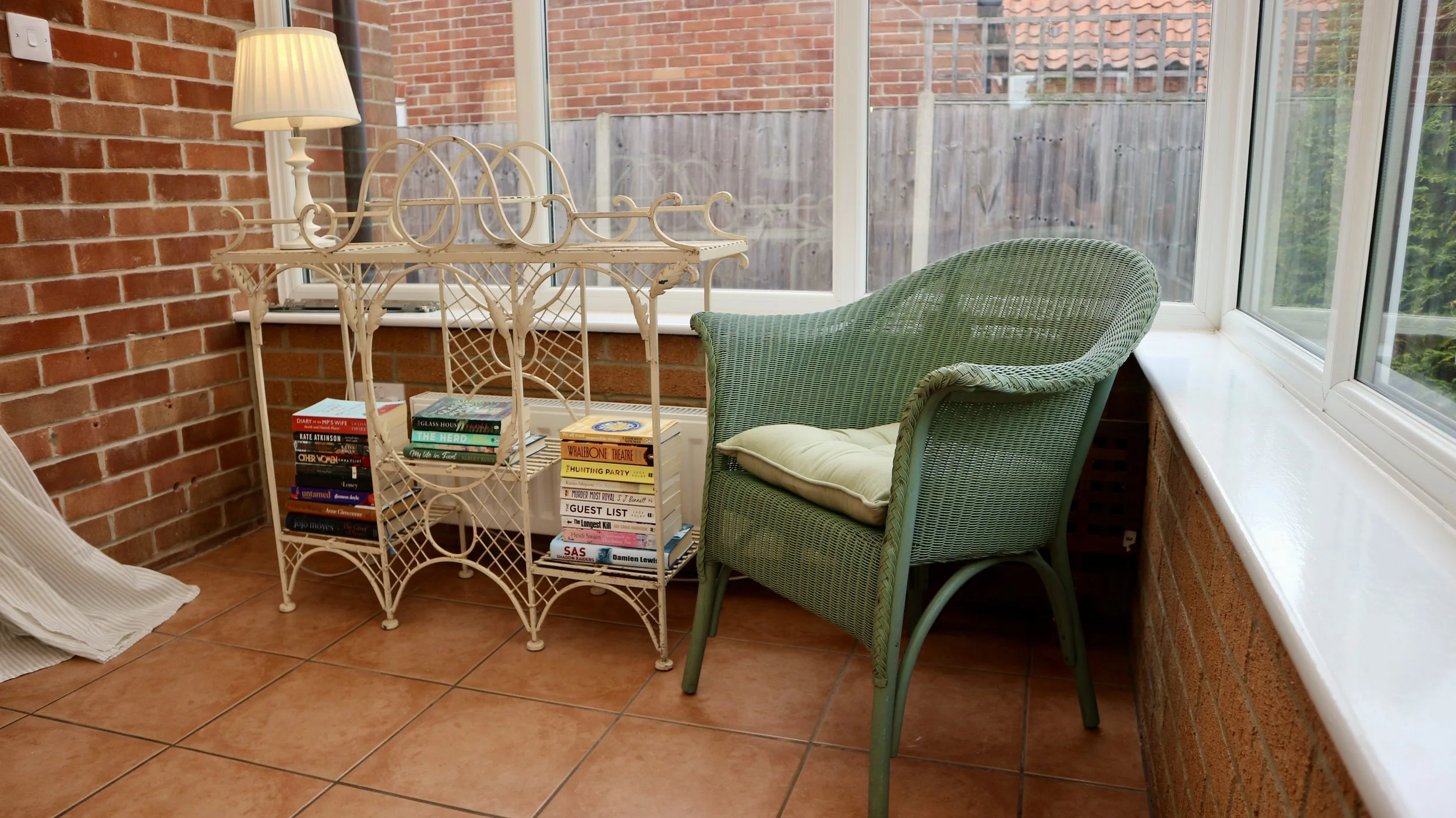 Conservatory with brick walls, tiled floor, a white wicker shelf with books, a green wicker armchair with a cushion, and large windows.