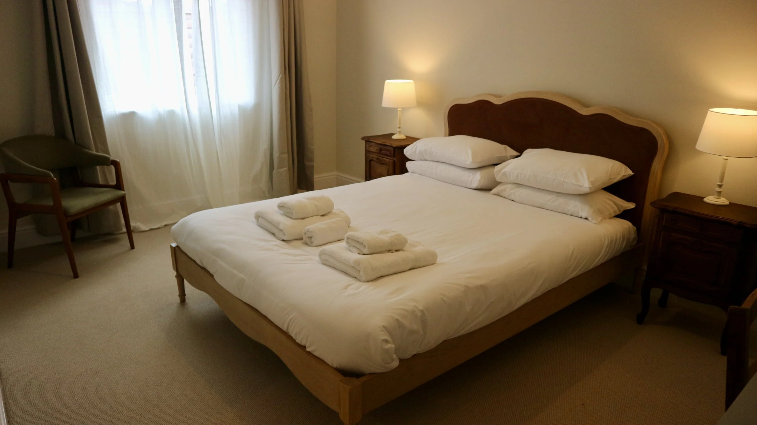 Master bedroom with a neatly made bed, four folded towels, two bedside lamps, a wooden side table, a chair, and curtains covering a window.