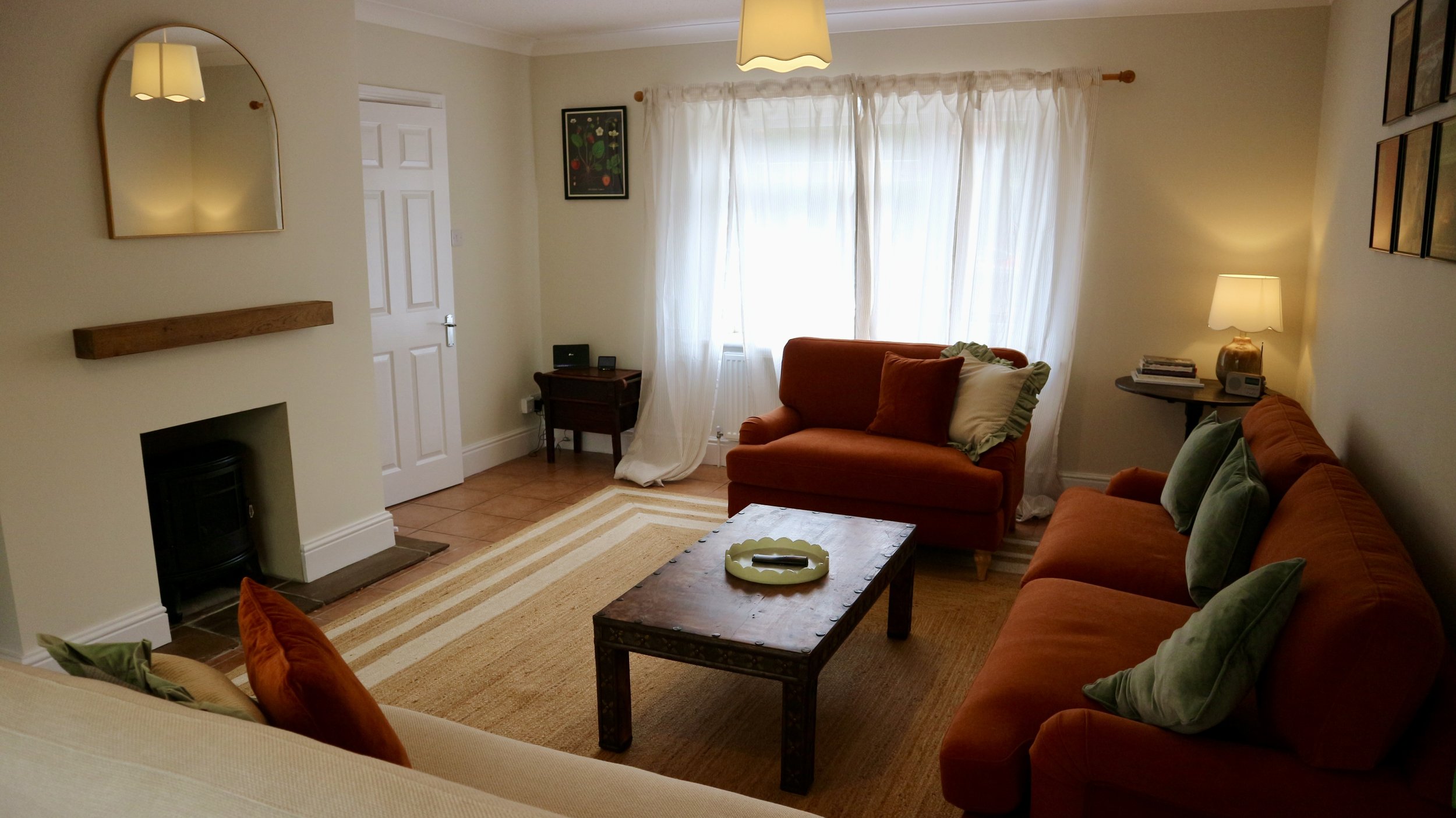 Living room with orange sofas, a wooden coffee table, cream walls, and white curtains covering a window. There are framed pictures on the wall, a mirror, and lamps providing warm lighting.
