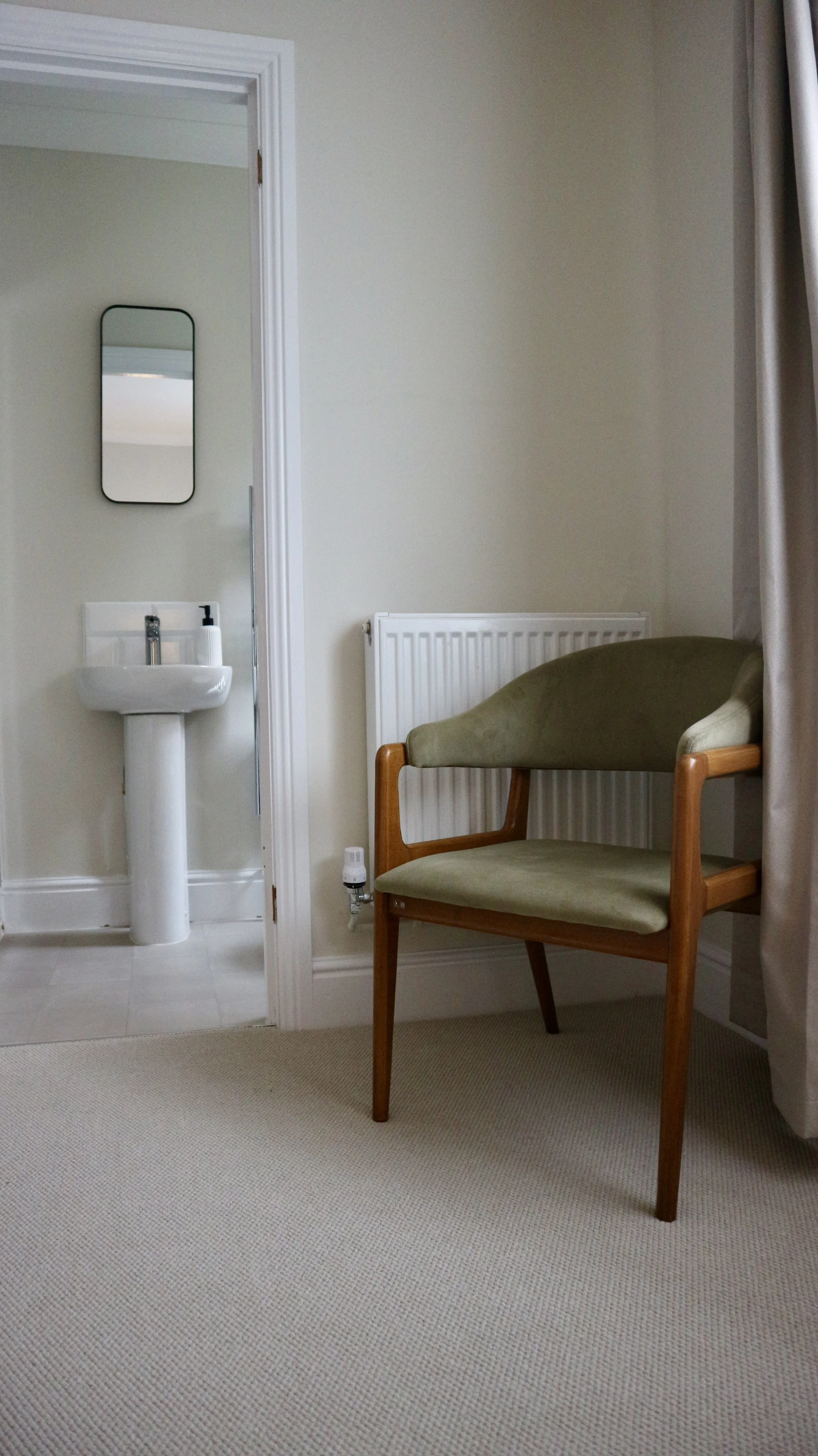 The master bedroon with a green upholstered armchair next to a white radiator, beige carpet, and a doorway leading to a small bathroom with a pedestal sink and a mirror.
