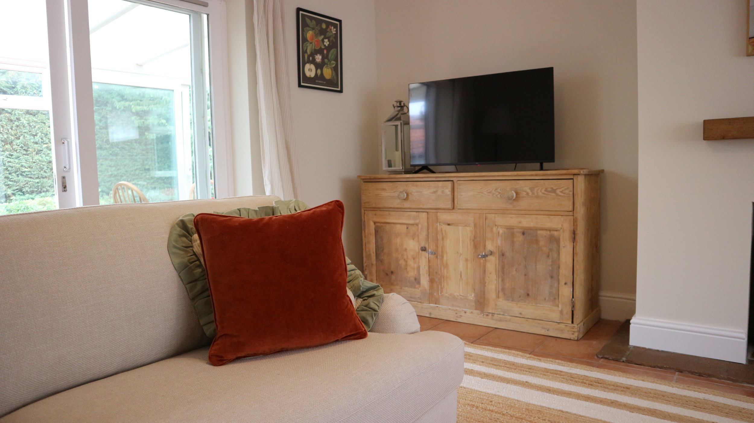 Living room with beige couch, rust-coloured pillow, wooden cabinet with TV, sliding glass doors leading outside, and framed floral artwork on the wall. Blackbird Cottage, recently refurbished.