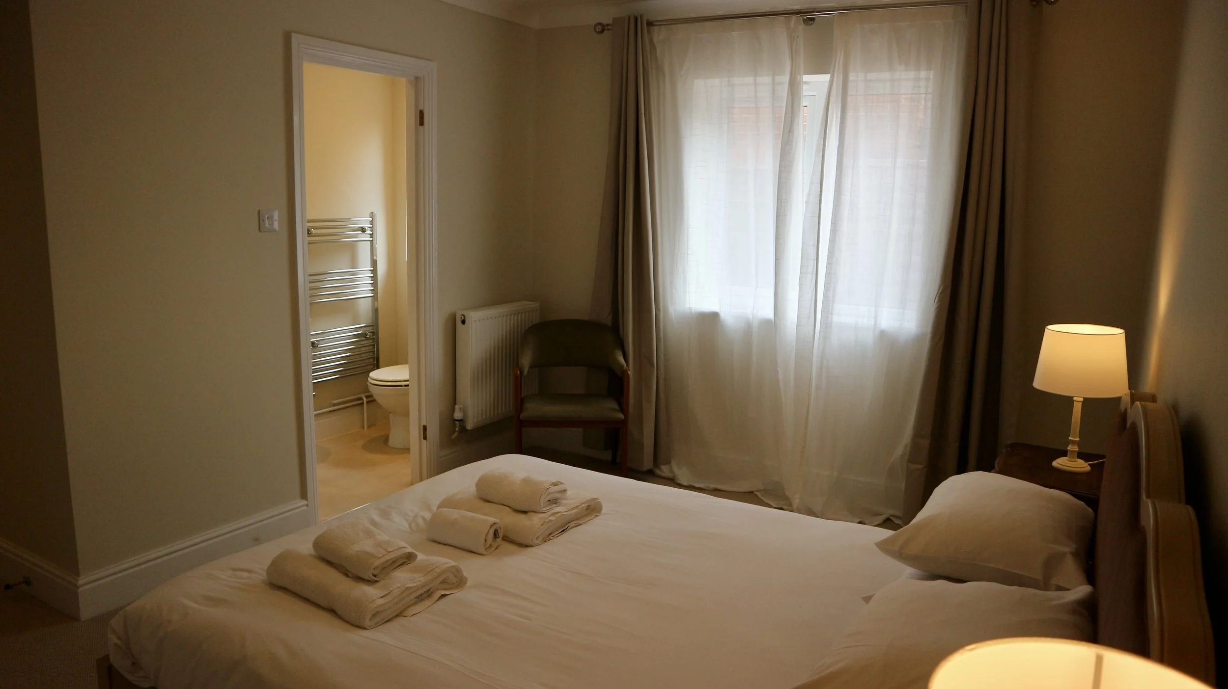 A cosy bedroom with a bed covered in white linens, four neatly folded towels on top, a bedside table with a lamp, a window with sheer curtains, a small chair, and a view into an en-suite bathroom with a toilet and a towel radiator.